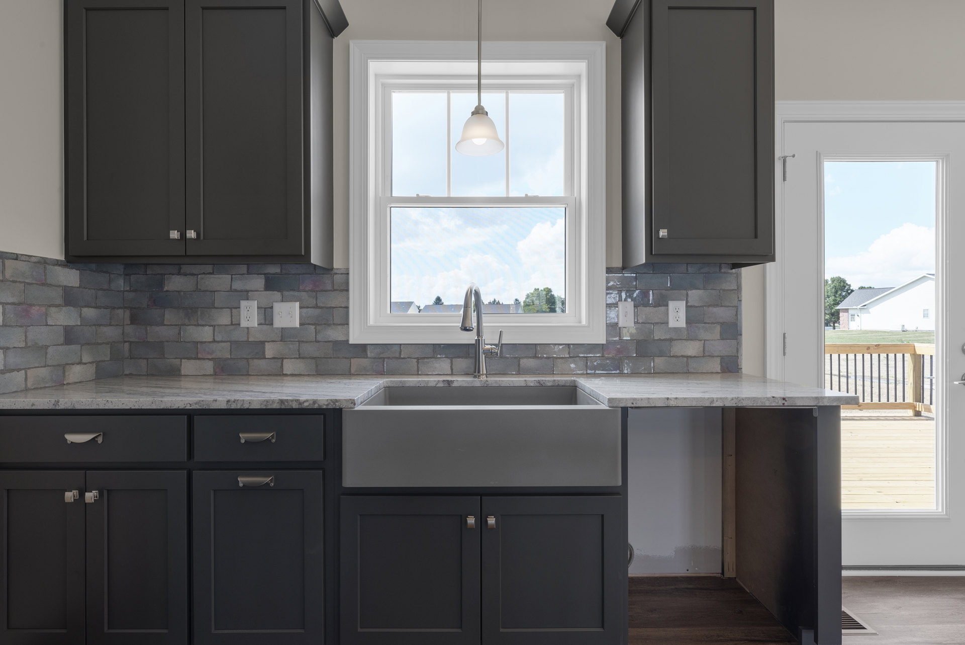 Grey kitchen cabinets with brushed metal handles, stone countertop, stainless steel sink and tap, brick accent wall with light switch, ceiling light fixture, large window letting