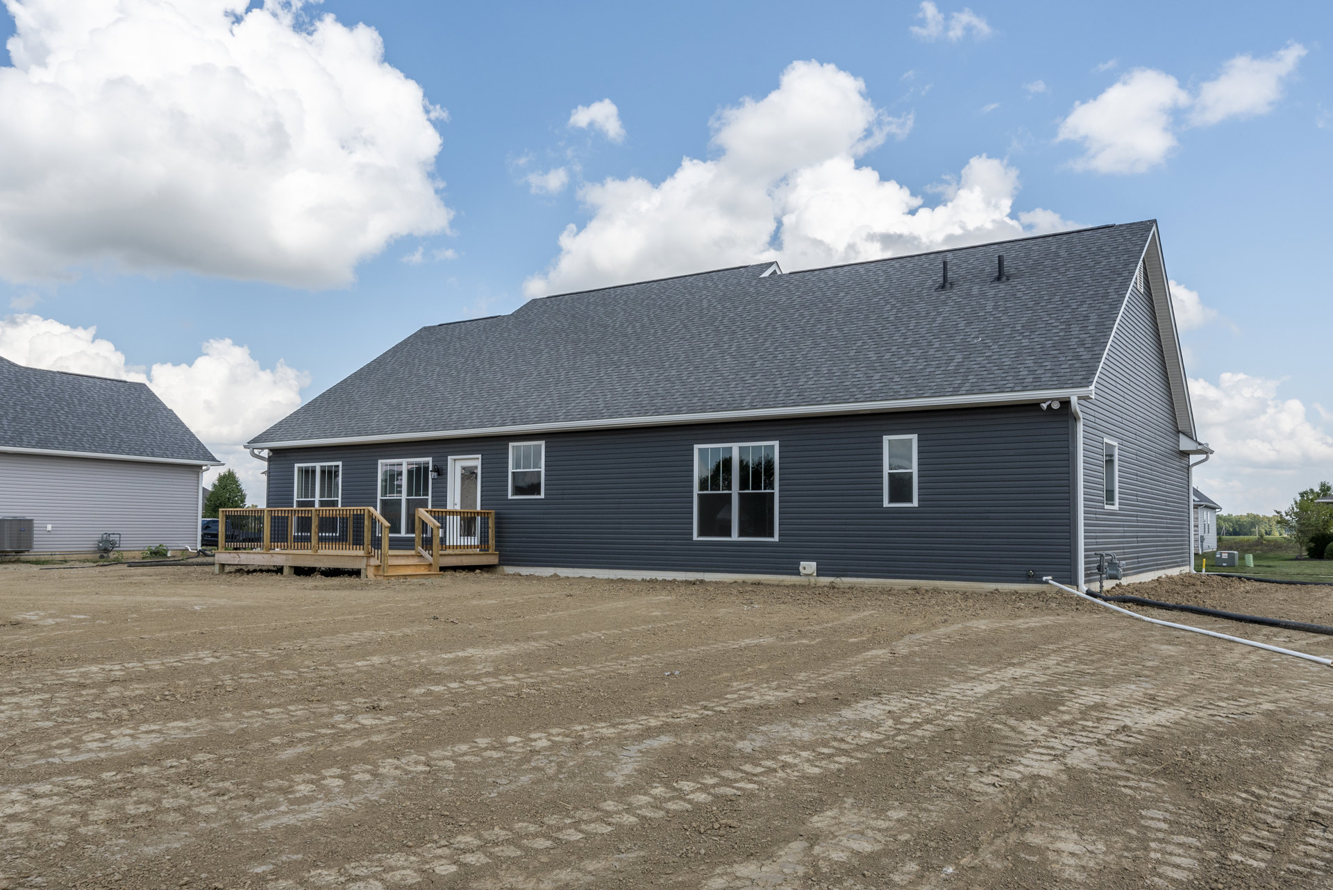 Two-story house with blue roof, white-trimmed windows, wooden deck featuring black railing, surrounded by a dirt field and tire-marked road, under a cloudy sky.