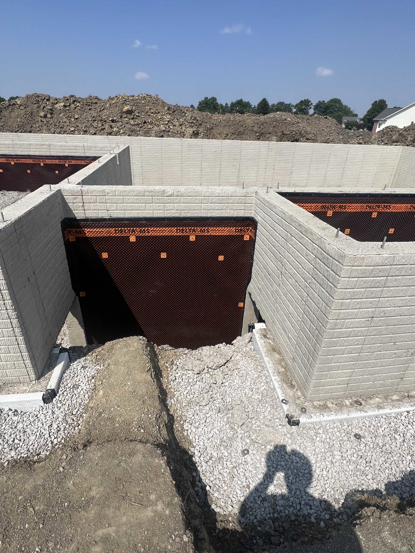 Concrete house foundation with a rectangular hole in the wall, brown and orange grid pattern on brick, pile of wood and rocks in foreground, blue sky with scattered clouds, trees