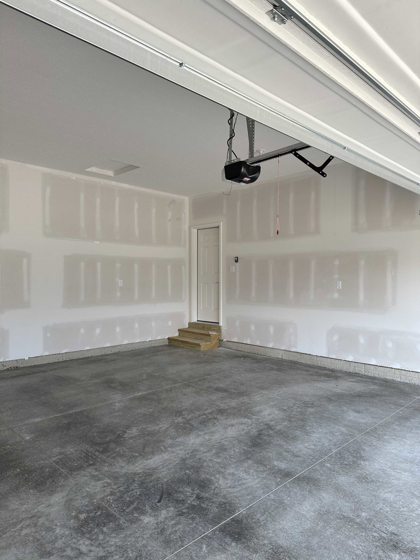 Garage interior featuring a concrete floor with a raised step in the corner, a vehicle lift, white door ajar, wooden staircase against a white plaster wall, overhead garage door