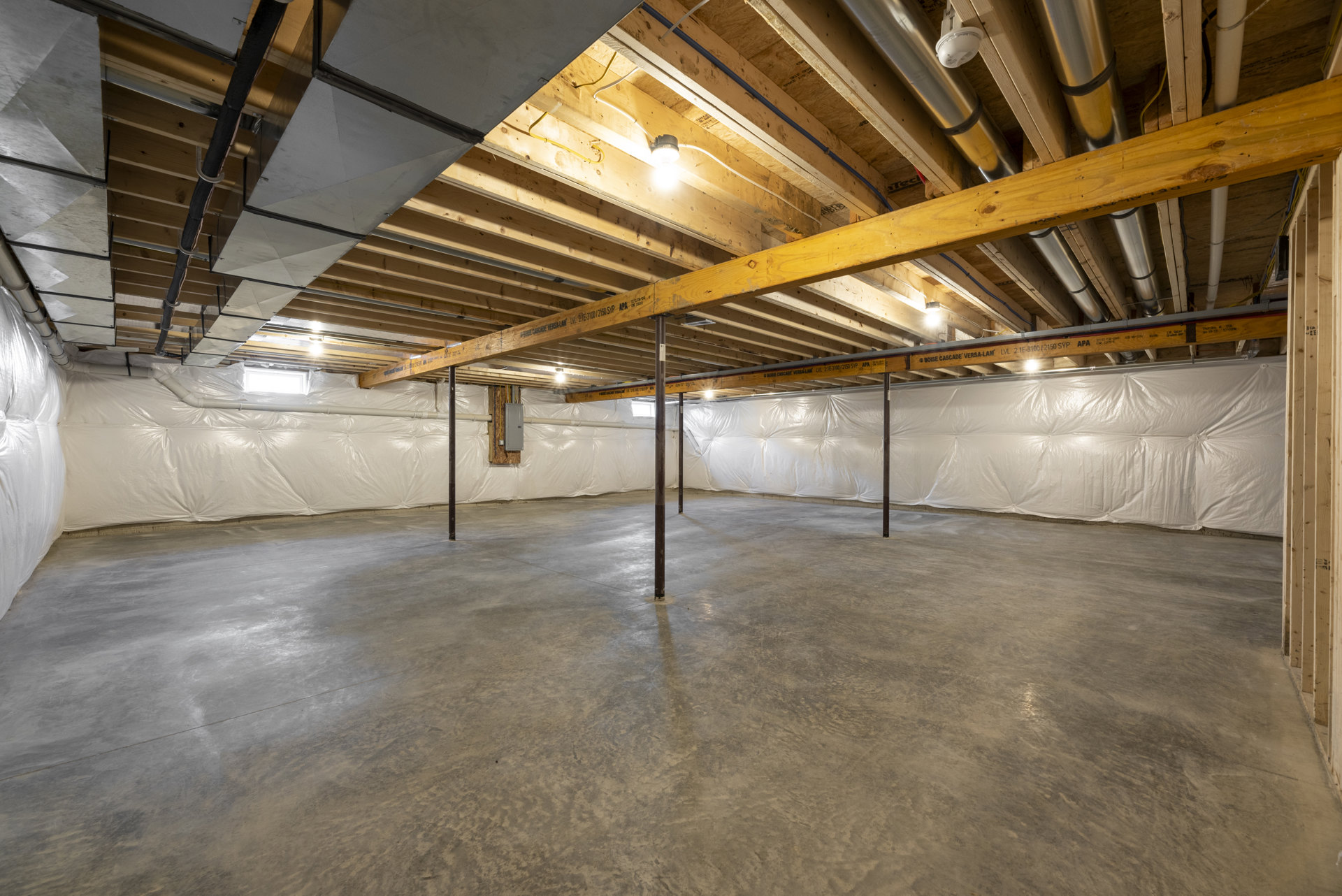 Concrete-floored basement room with exposed steel beams, visible ceiling pipes, and building insulation along the walls