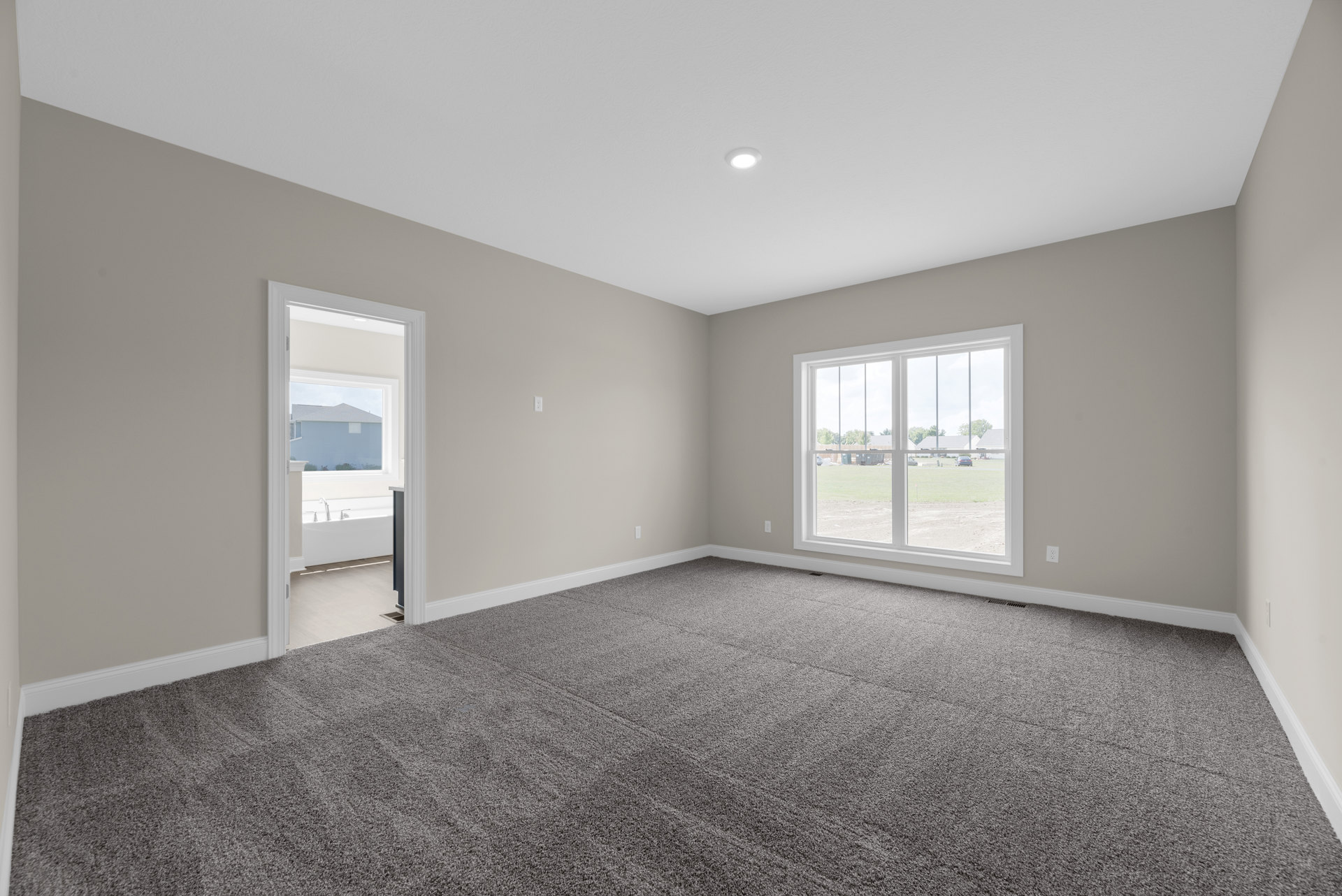 Bright room with grey carpeted floor, white walls, large window overlooking field and parked cars