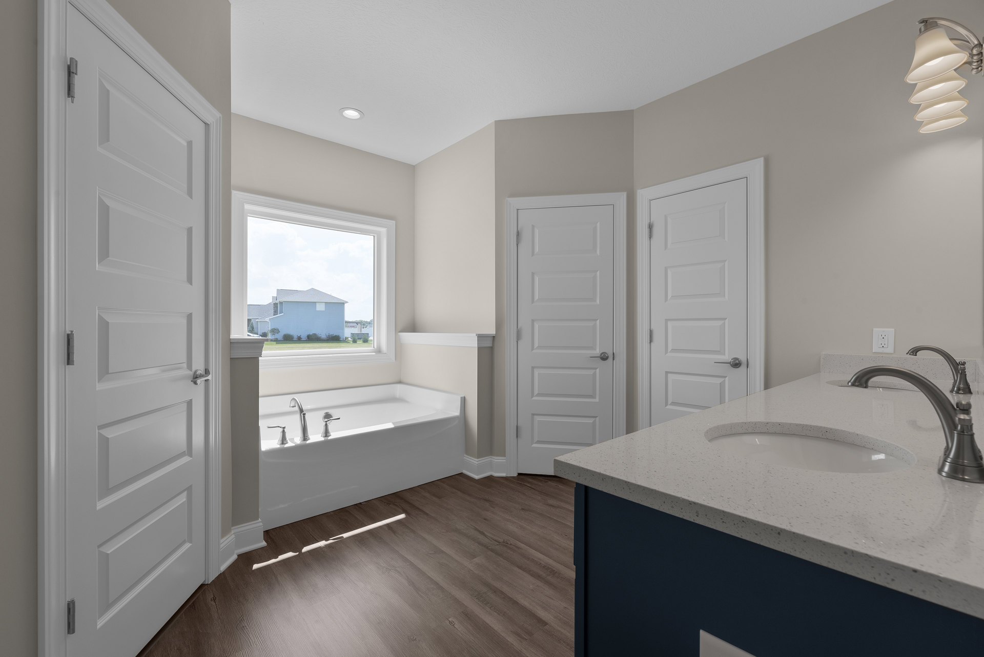 Freestanding bathtub with chrome faucets on wood floor, large window with white trim, bathroom vanity with sink and silver faucet, white door with silver handle, ceiling pendant