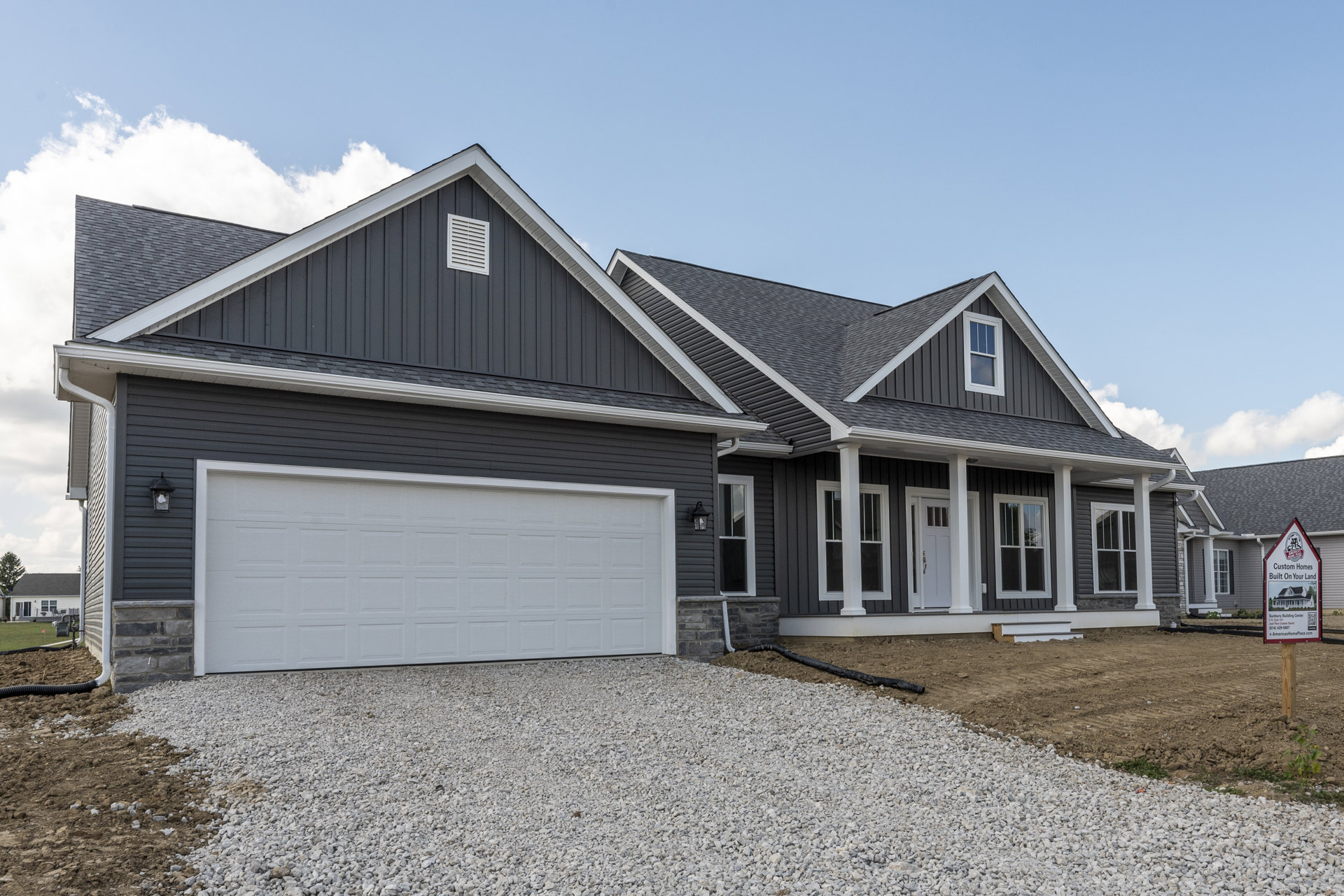 Two-story home with white siding, black roof, white garage door, gravel driveway, white wall vent, and a window; sign with house illustration near entrance.