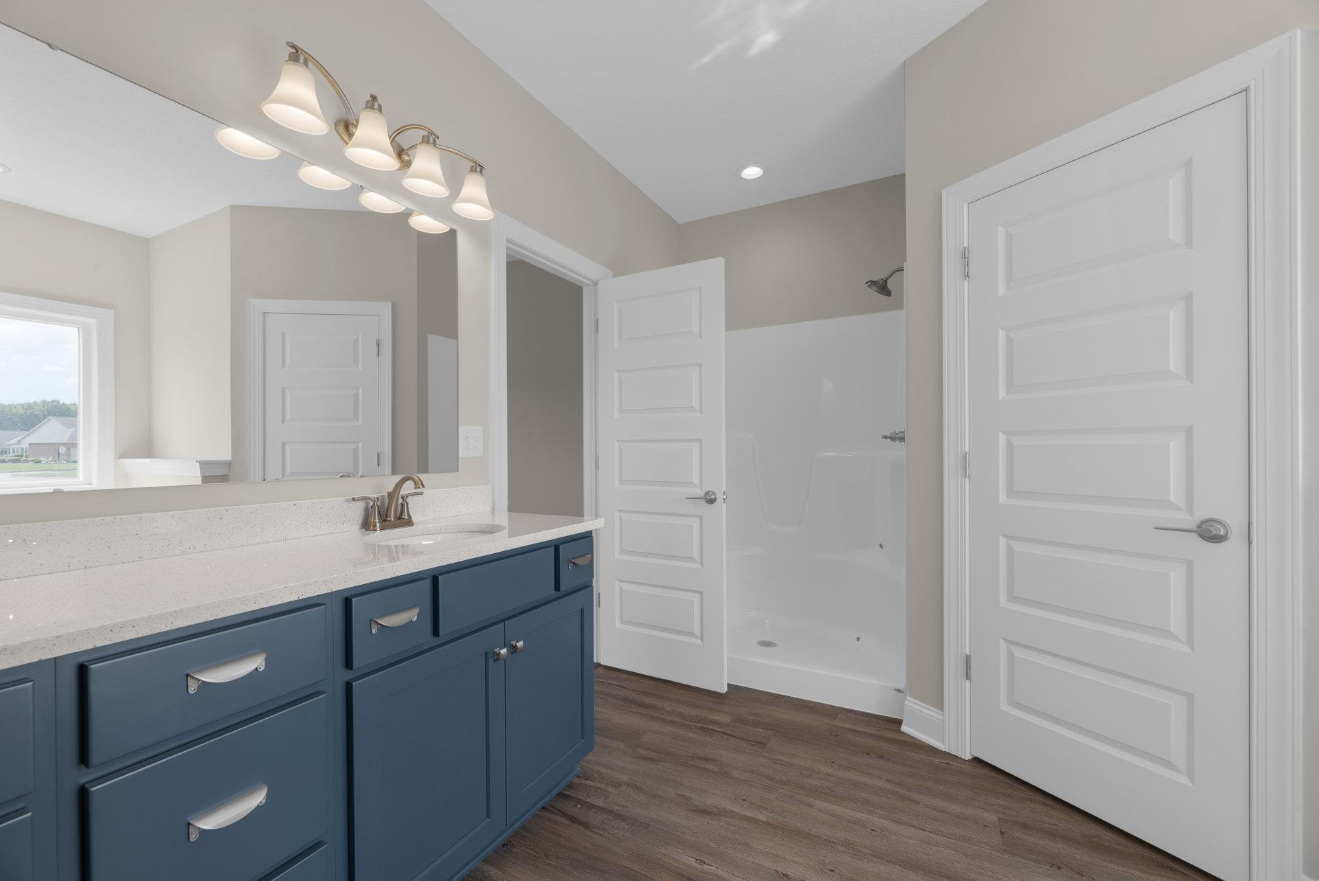 Bathroom with blue cabinetry and silver handles, white sink, glass shower enclosure, white door with silver handle, light fixture on ceiling, neutral wall and floor finishes