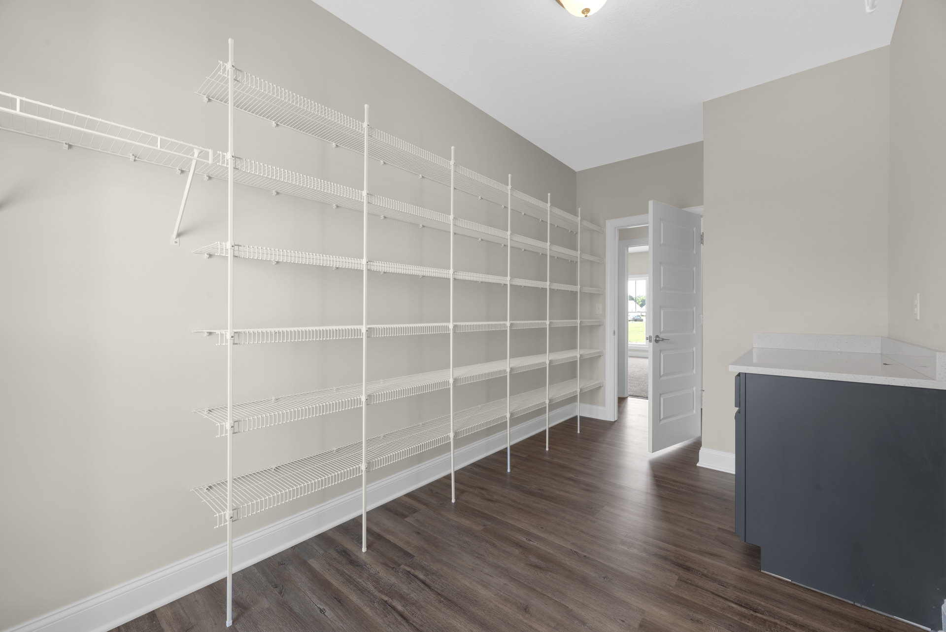 Built-in white shelves along a plaster wall, light-colored countertop, white door with silver handle, hardwood floor, and smooth ceiling in a modern kitchen.
