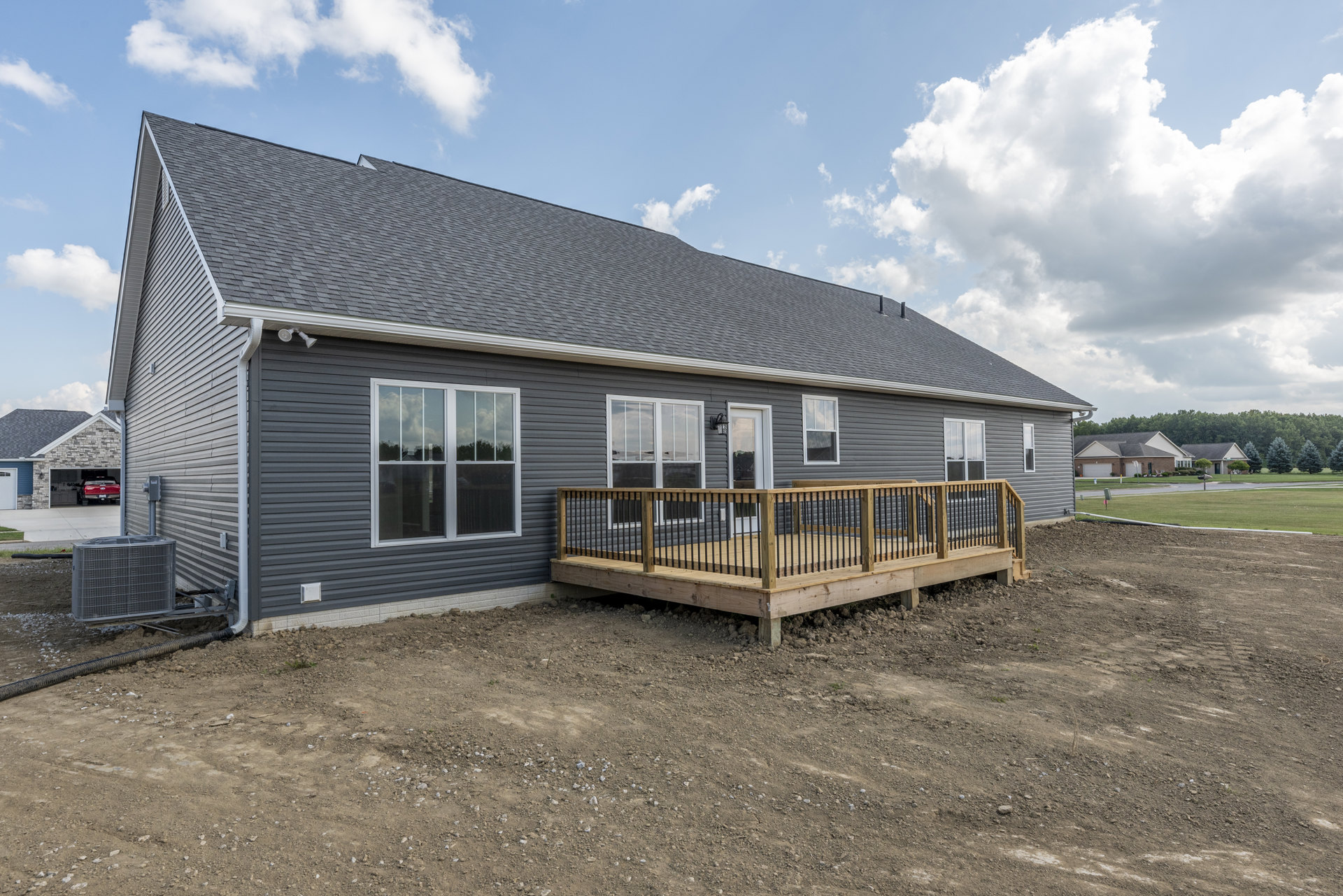 Wooden deck with metal railing attached to a house, white-framed window, large metal utility box with grid, dirt yard, cloudy sky above