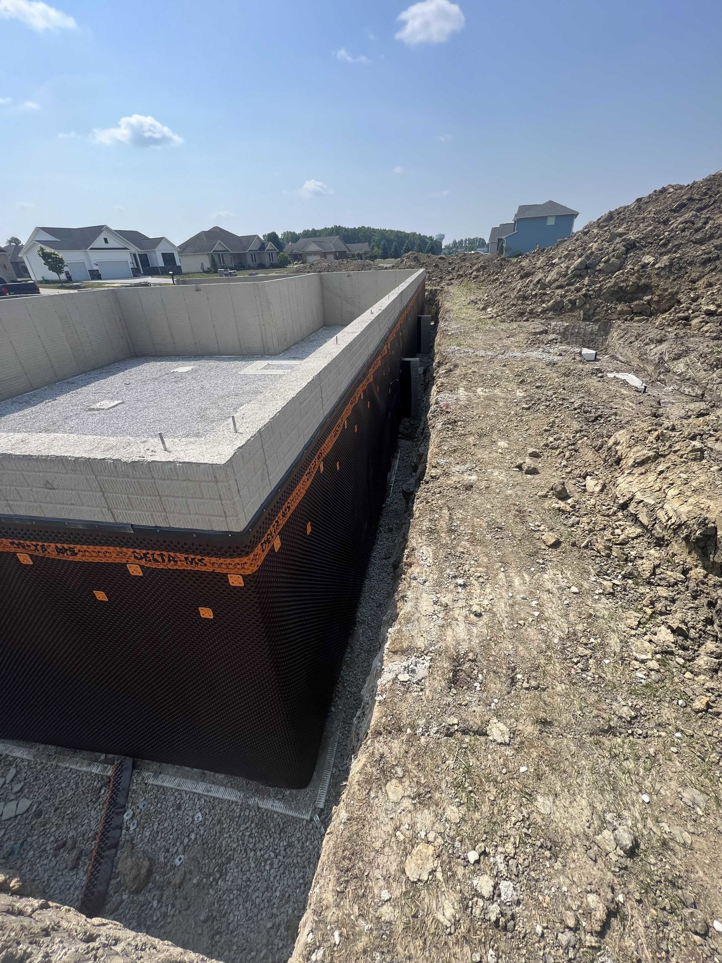 Concrete house foundation surrounded by dirt and rocks, black and white retaining wall, blue sky with clouds, trees in background