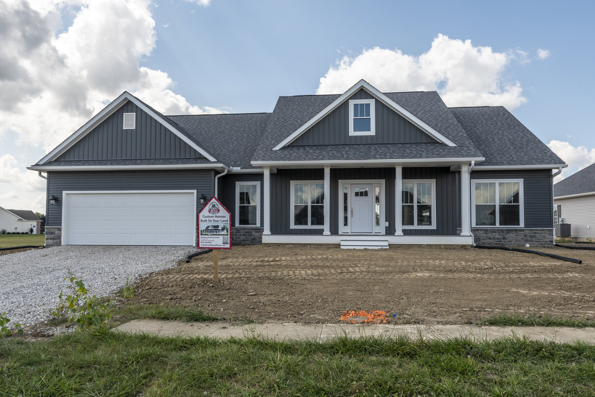 White two-story home with black trim, white garage door, glass-paneled front door, large windows reflecting clouds, and real estate sign on landscaped front lawn.