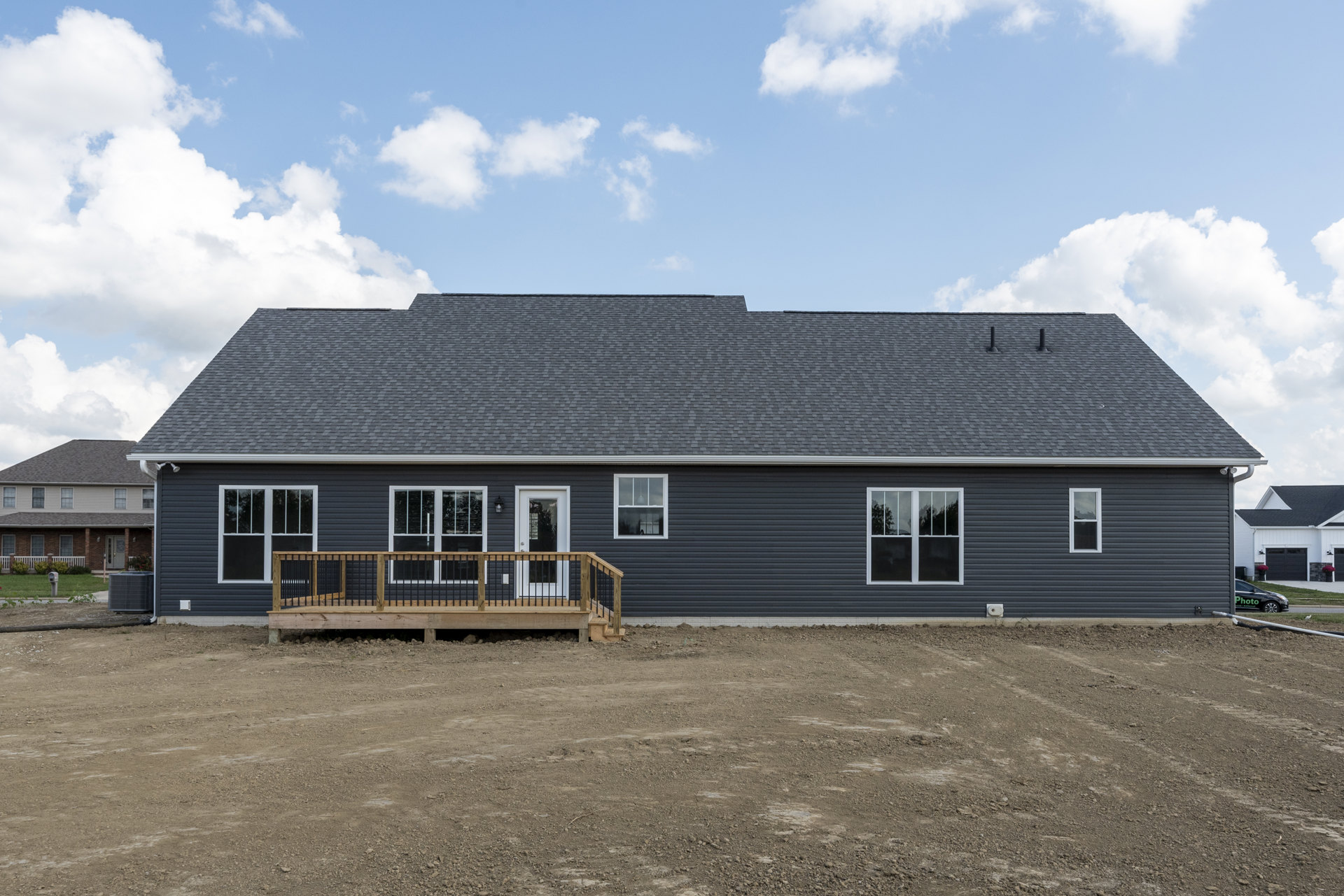 Wooden deck with black railing attached to a house, white-framed windows, porch area, dirt yard in foreground, cloudy sky above