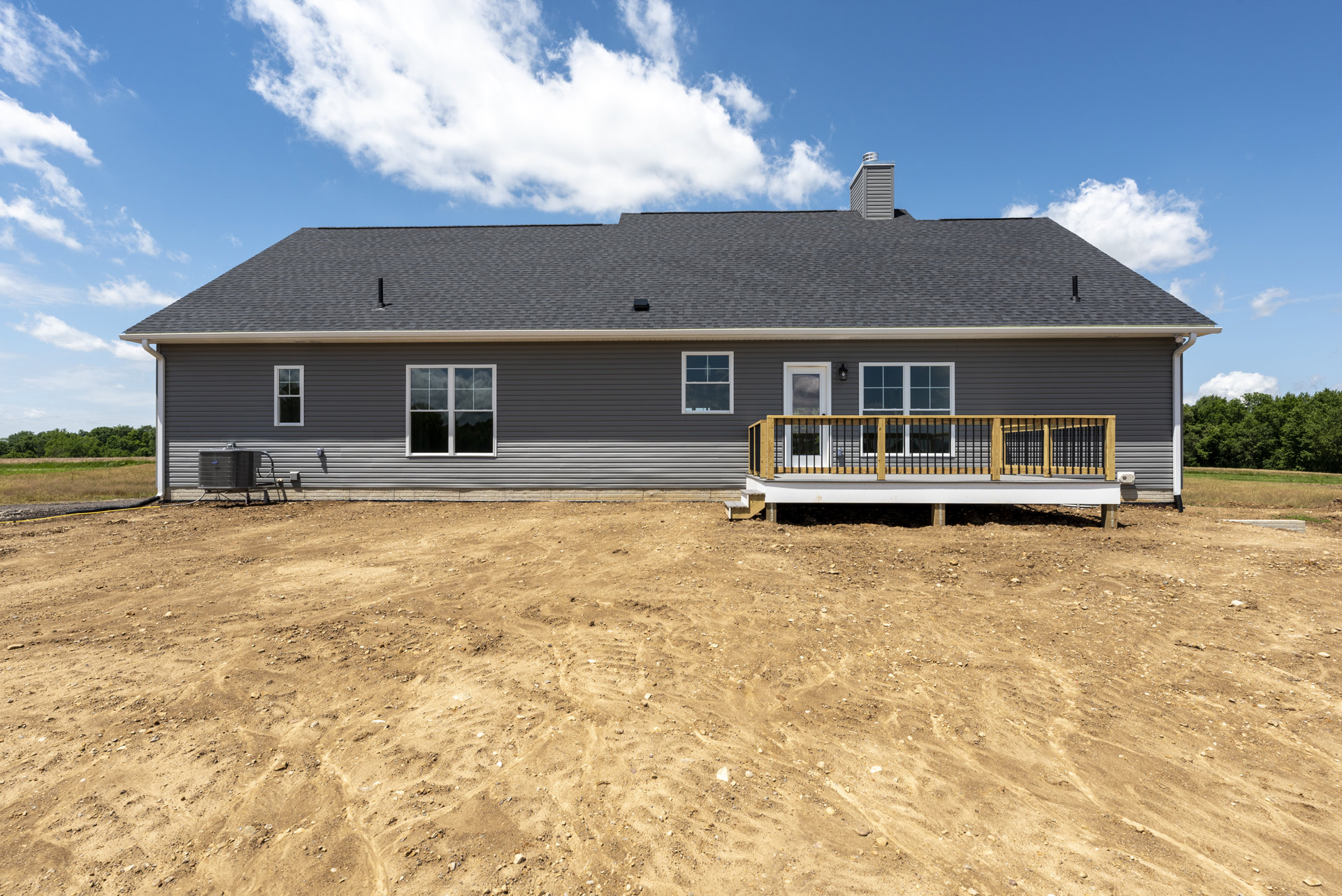Two-story house with white-framed windows, wooden deck and railing, dirt yard, metal fence, and trees under a blue sky
