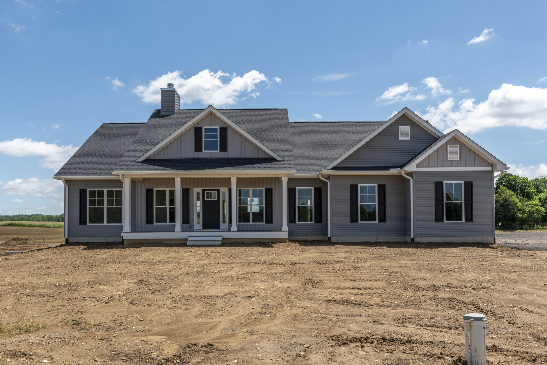 Two-story house with white siding, black shutters, and white-framed windows, front porch with white columns, surrounded by a dirt field under a cloudy sky