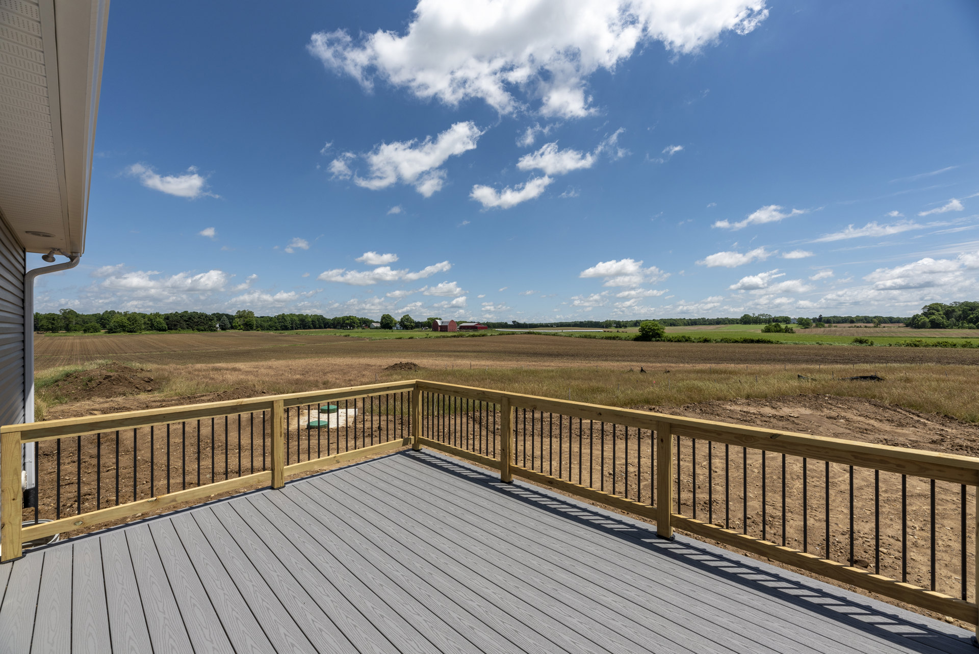 Wood deck bordered by a horizontal plank fence, overlooking a grassy field under a blue sky with scattered clouds; rain gutter attached to house, green plastic lids resting on dirt