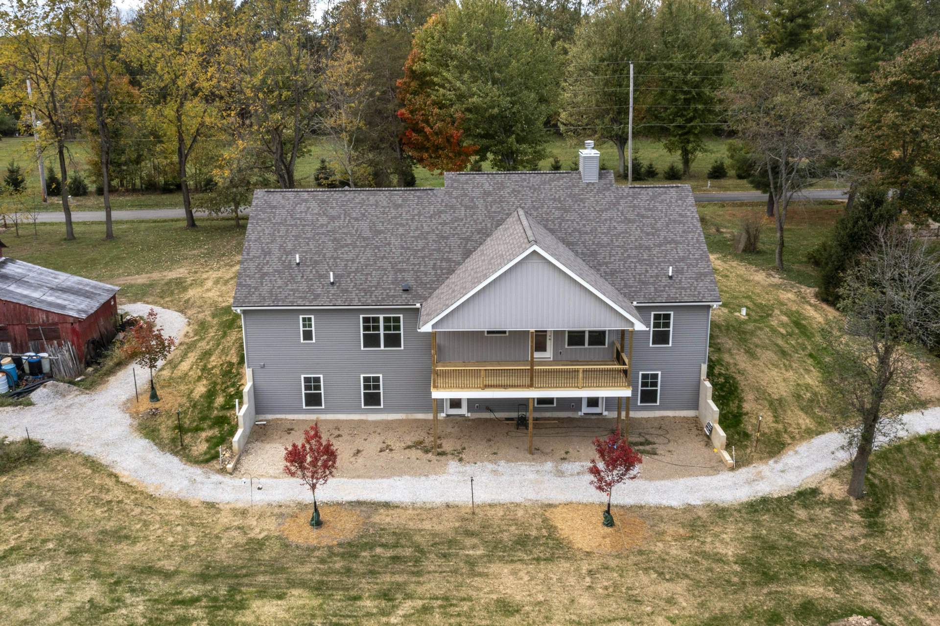 Two-story house with gray siding, multi-pane windows, elevated deck, covered porch, concrete driveway, landscaped yard, and small tree with red leaves.