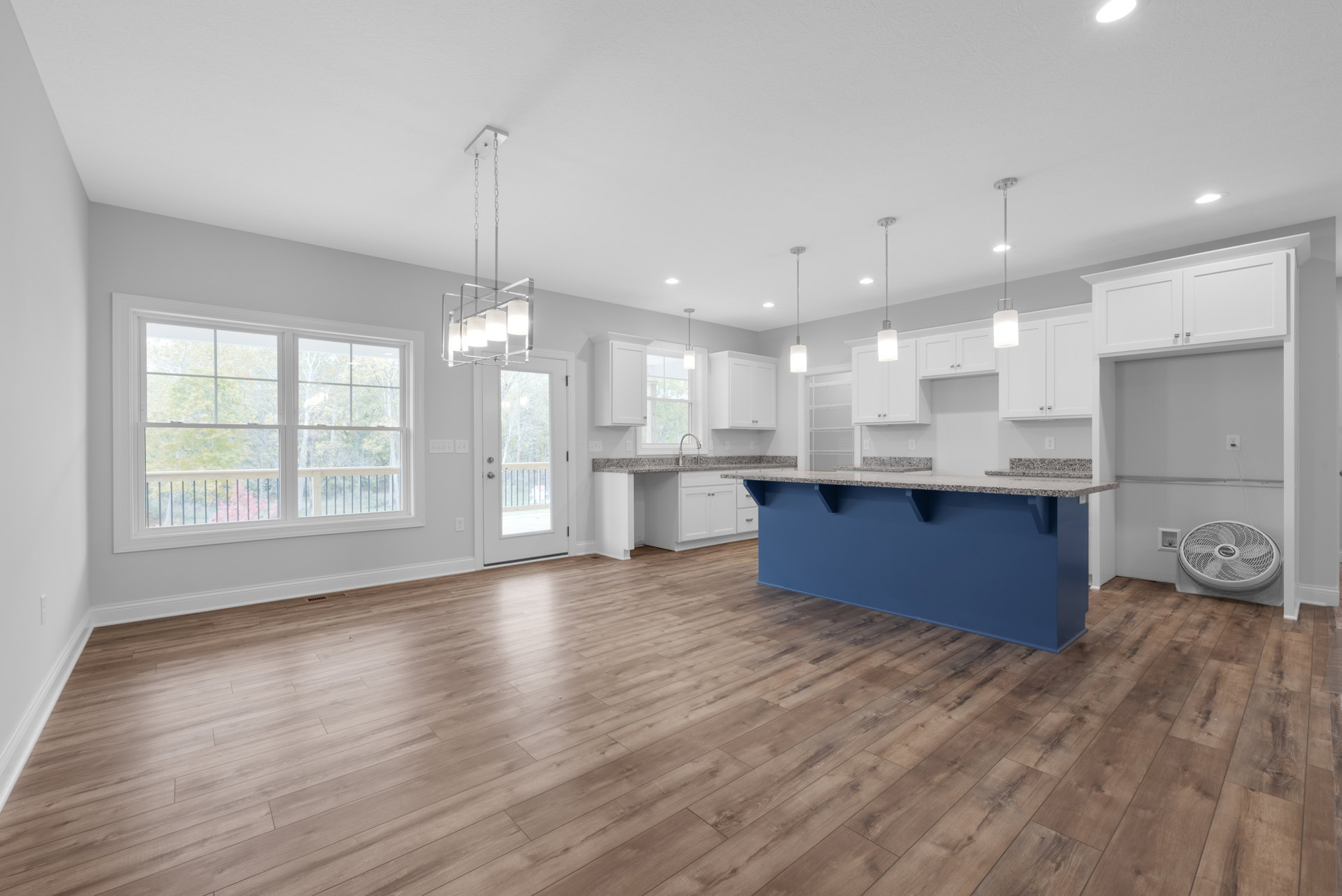 Blue kitchen island with white countertops, wood flooring, large window overlooking trees with railing, white ceiling fan, white cabinetry, and glass door leading to outdoor area.