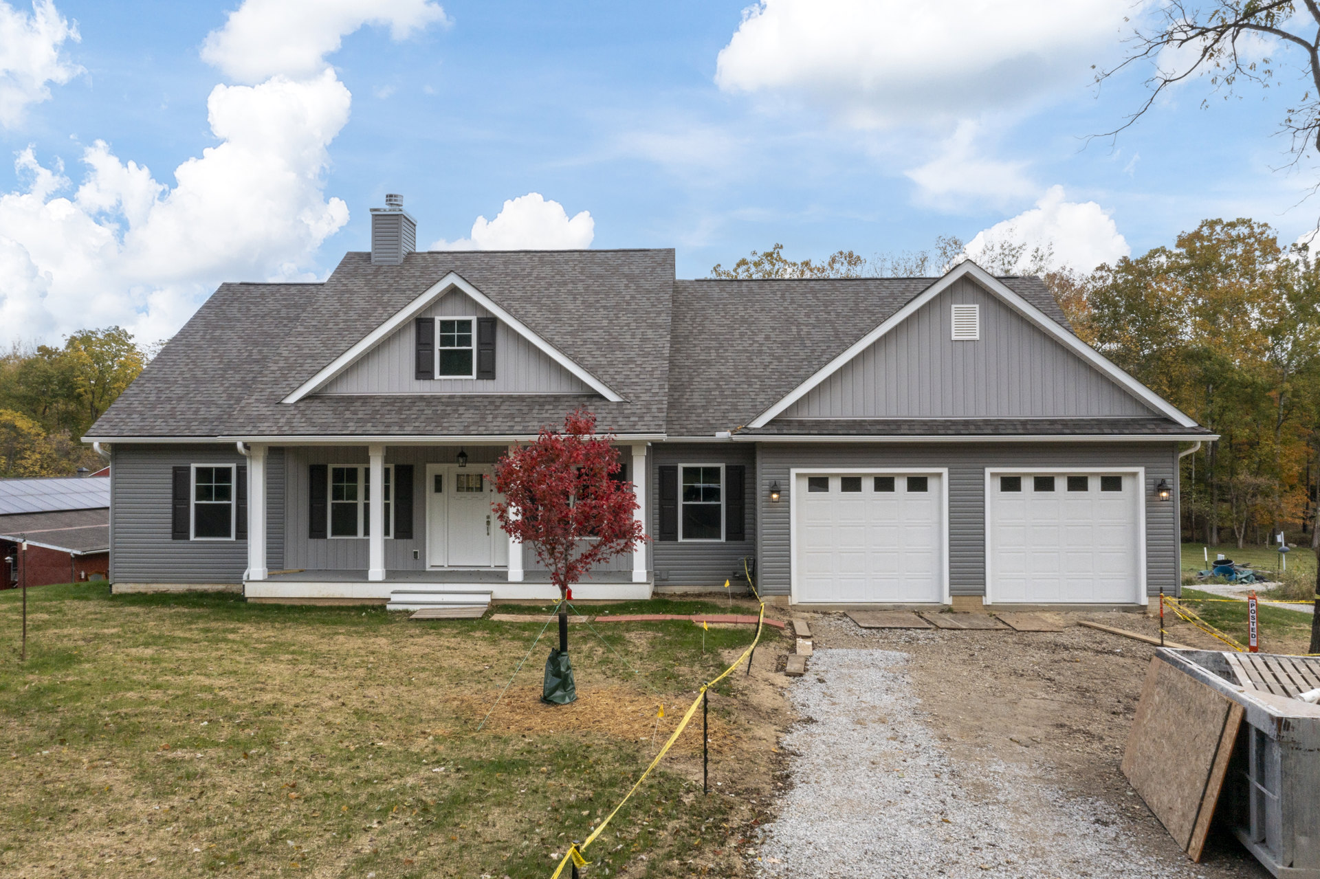 Two-story house with white garage door and multi-pane windows, red-leafed tree and dumpster in driveway, cloudy sky overhead