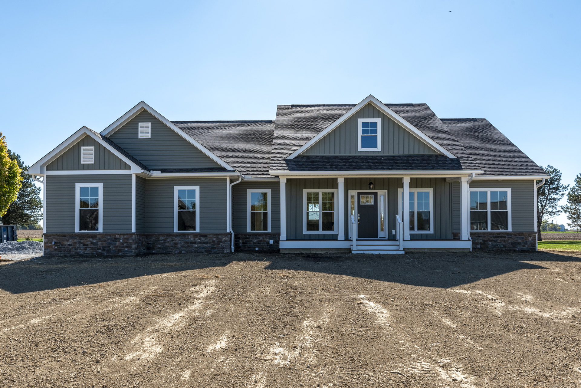 Two-story house with white porch railing, dirt driveway, large windows reflecting trees and sky, surrounded by grass and mature trees