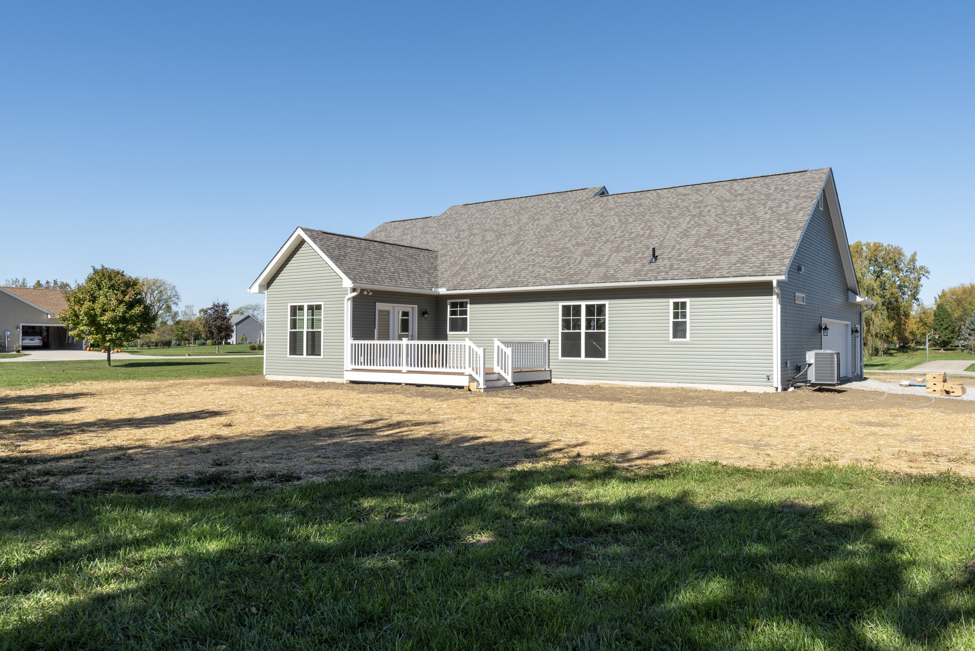 Two-story house with gray siding, white porch railing, large windows with white frames, shingled roof, wooden deck, and manicured green lawn surrounded by trees.