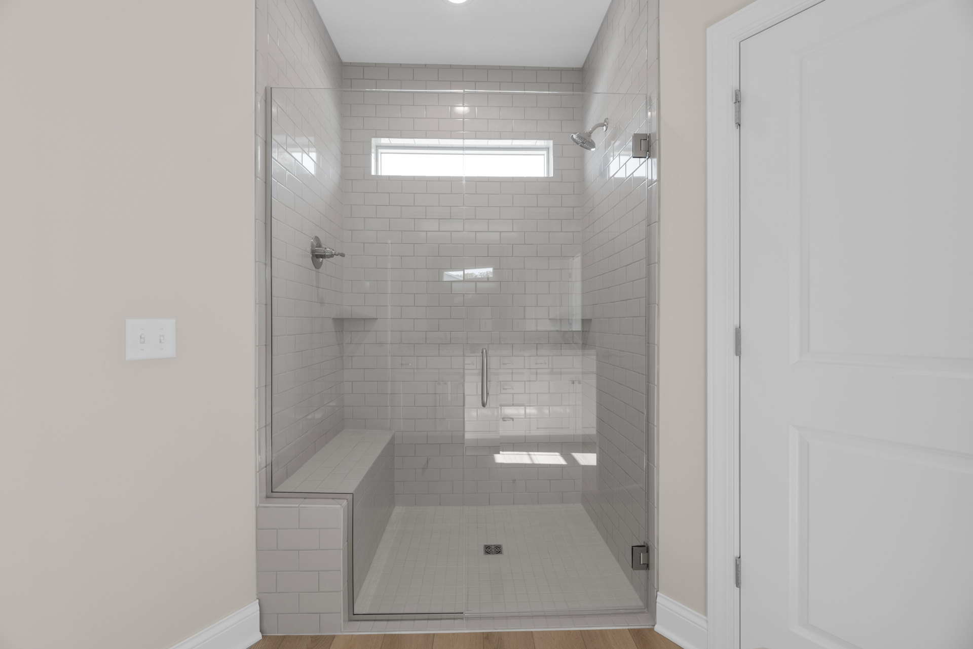 Glass shower enclosure with built-in bench, white door with metal hinge, tile floor featuring shower drain, close-up window, plaster walls, and dual light switches.