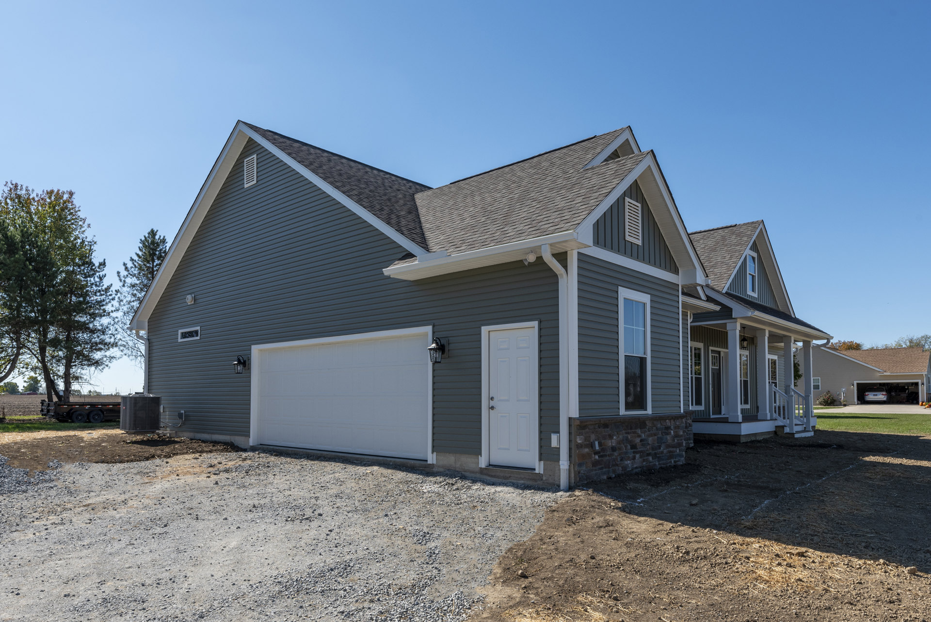 Two-story house with white siding, gray shingle roof, attached white garage door, silver handle, front porch, large window, black heat pump unit, and leafless tree in yard