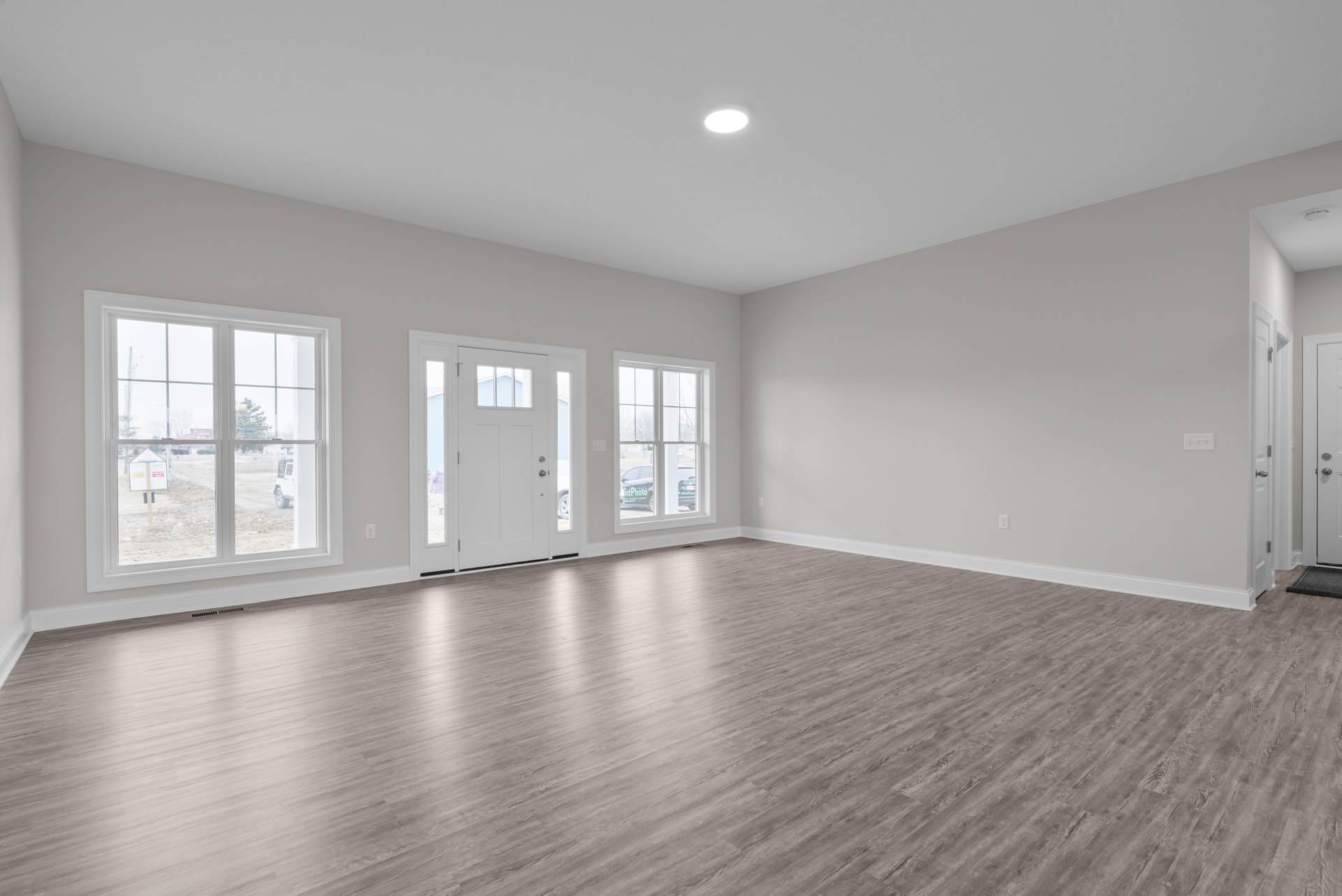 Room with wood flooring, white plaster walls, recessed ceiling light, multiple windows including one with a car visible outside, and a white door with glass panels.