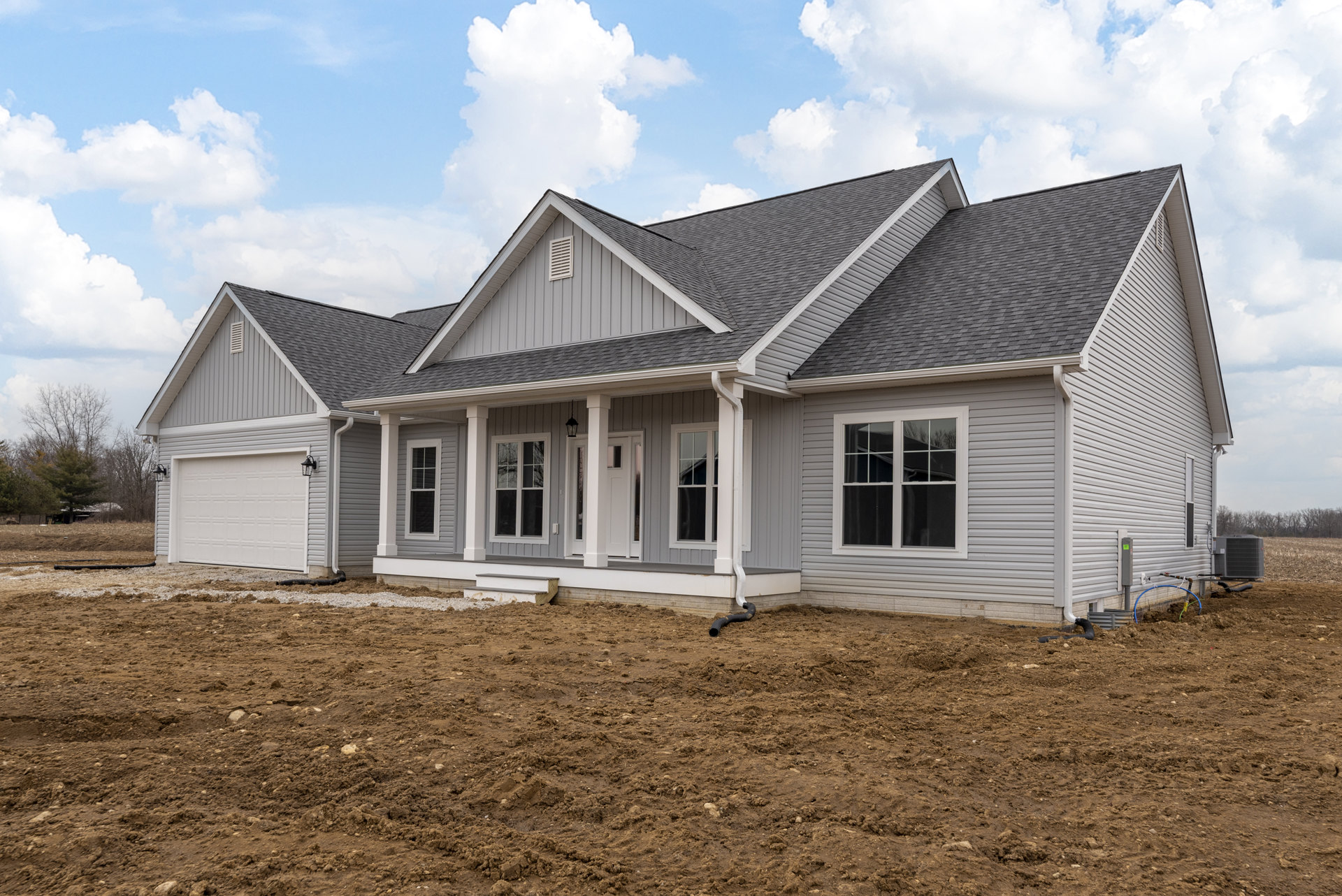 Two-story house with gray siding, white-framed windows, and a covered porch with a white door, set on a dirt lot with a black pipe near the foundation; Robert Frost Farm visible in