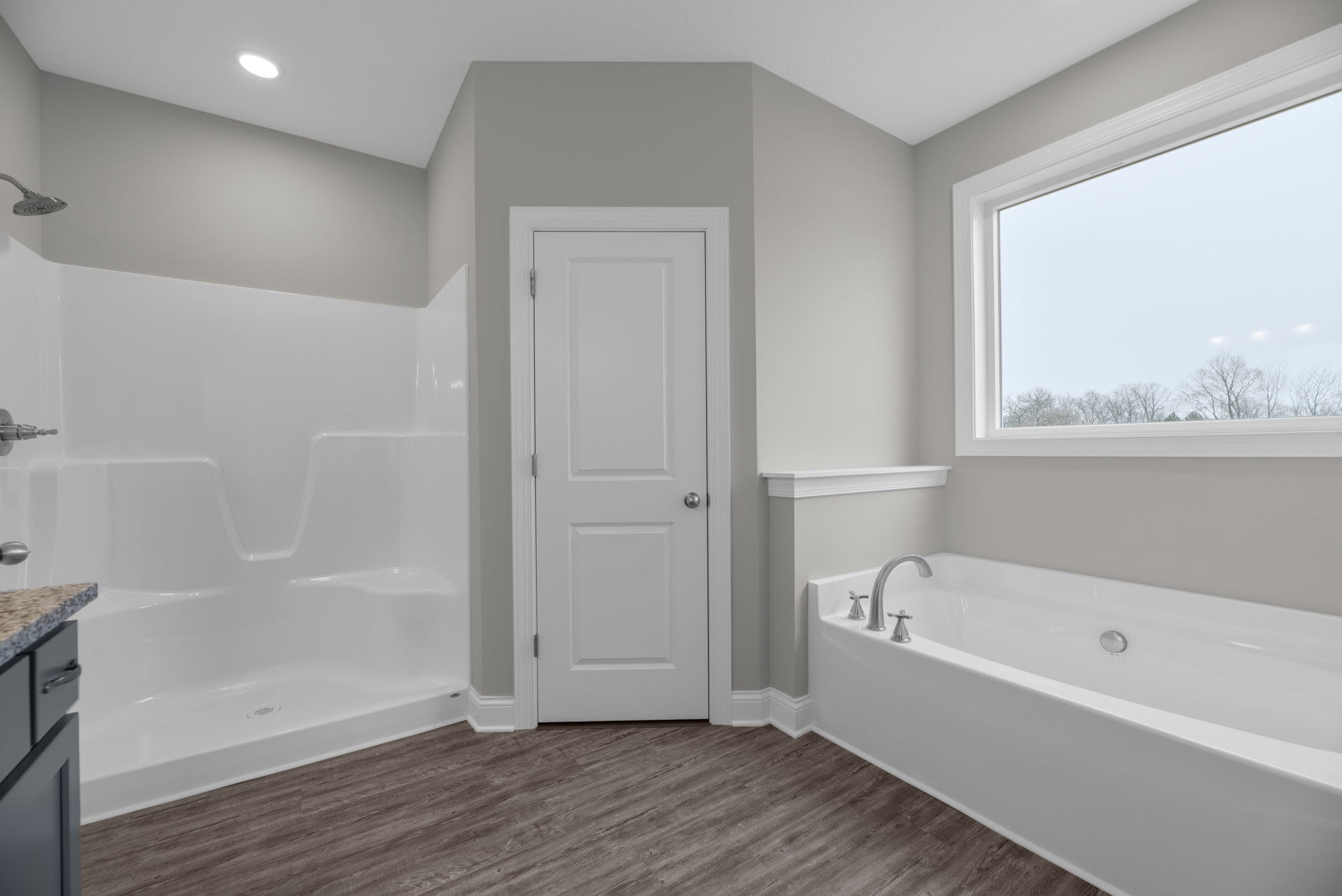 Freestanding white bathtub beneath a large window with outdoor trees visible, wood flooring, white door with silver knob, ceiling light fixture