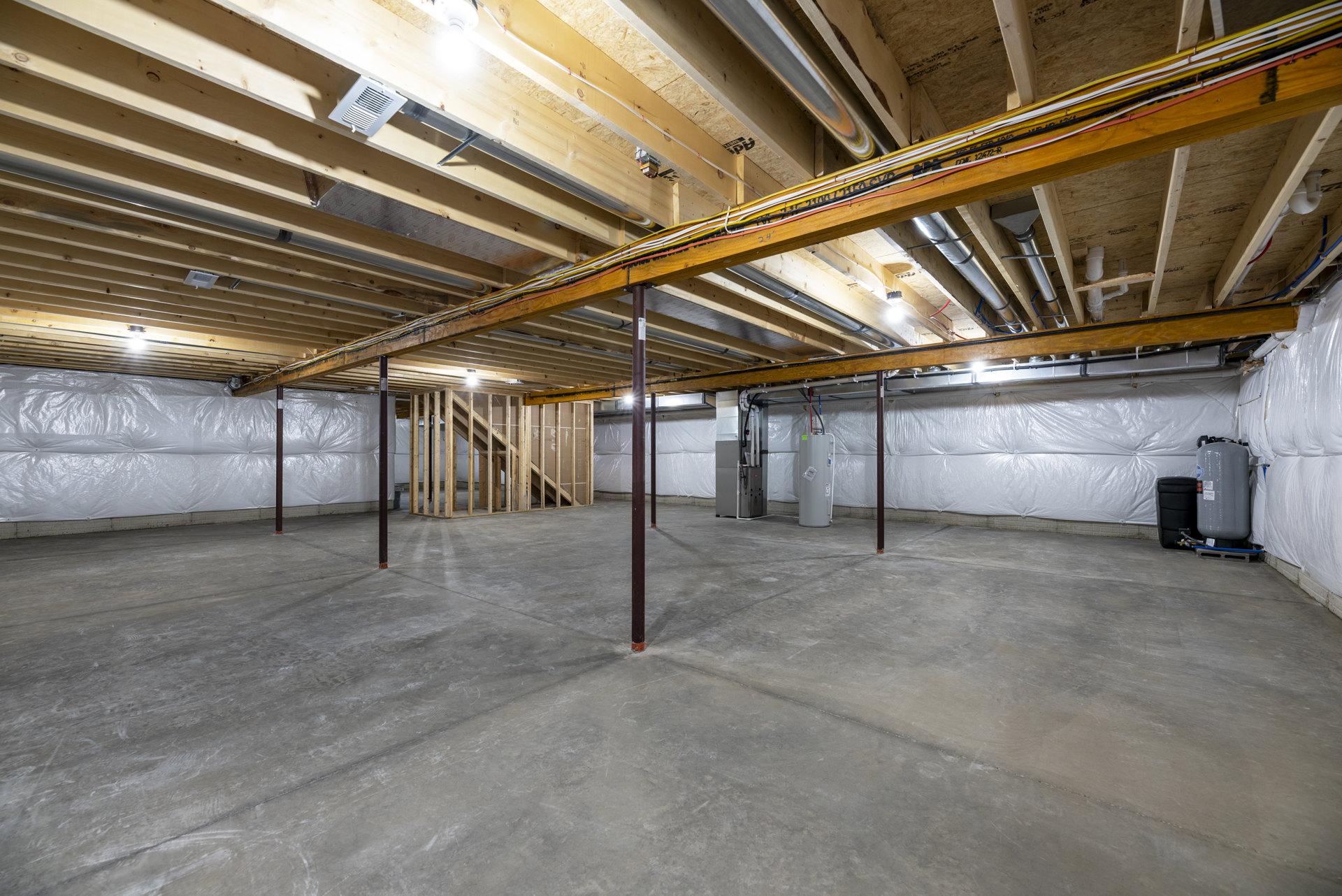 Unfinished basement with exposed wood and metal beams, concrete floor, white cylindrical container with warning sign, visible electrical wires and black cord.