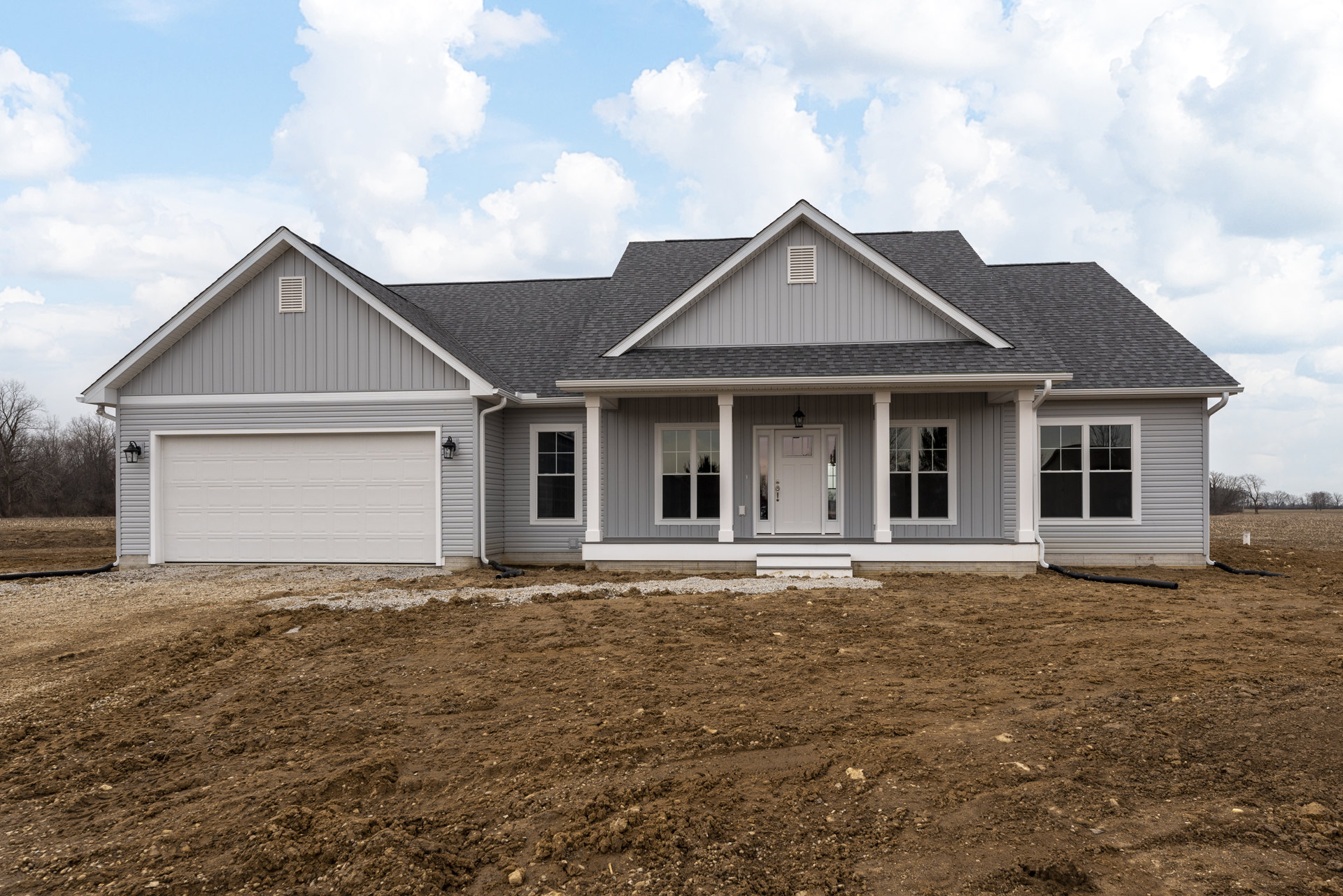 Two-story house with white siding, grey roof, white garage door, covered porch, and large windows reflecting trees, set behind a dirt field under a cloudy sky