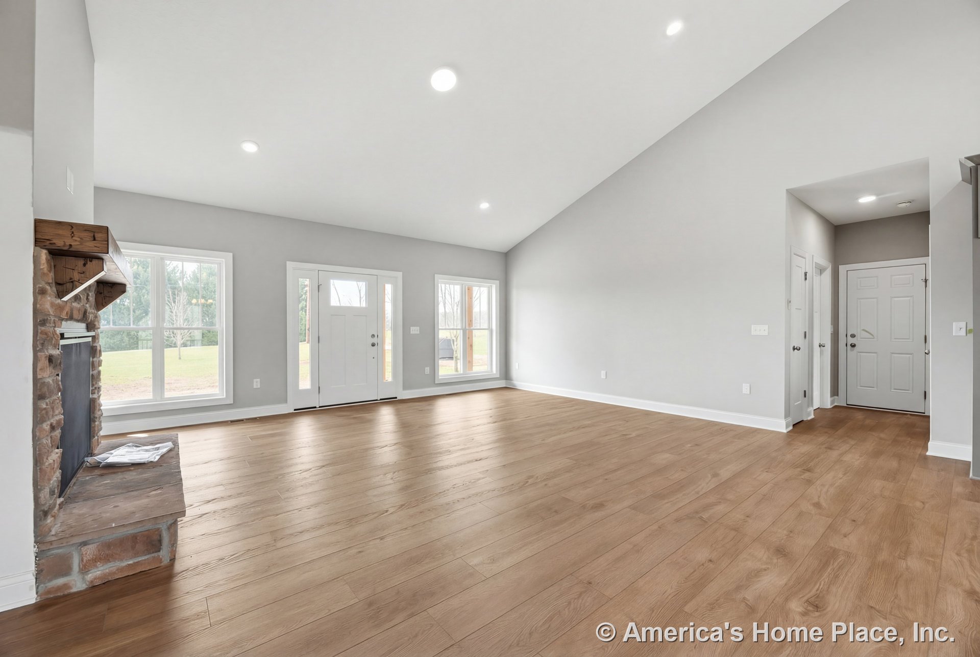 Vaulted ceiling living room with large windows, brick fireplace topped by wood mantel, light wood plank flooring, white trim, recessed lighting, and open layout with entry door.