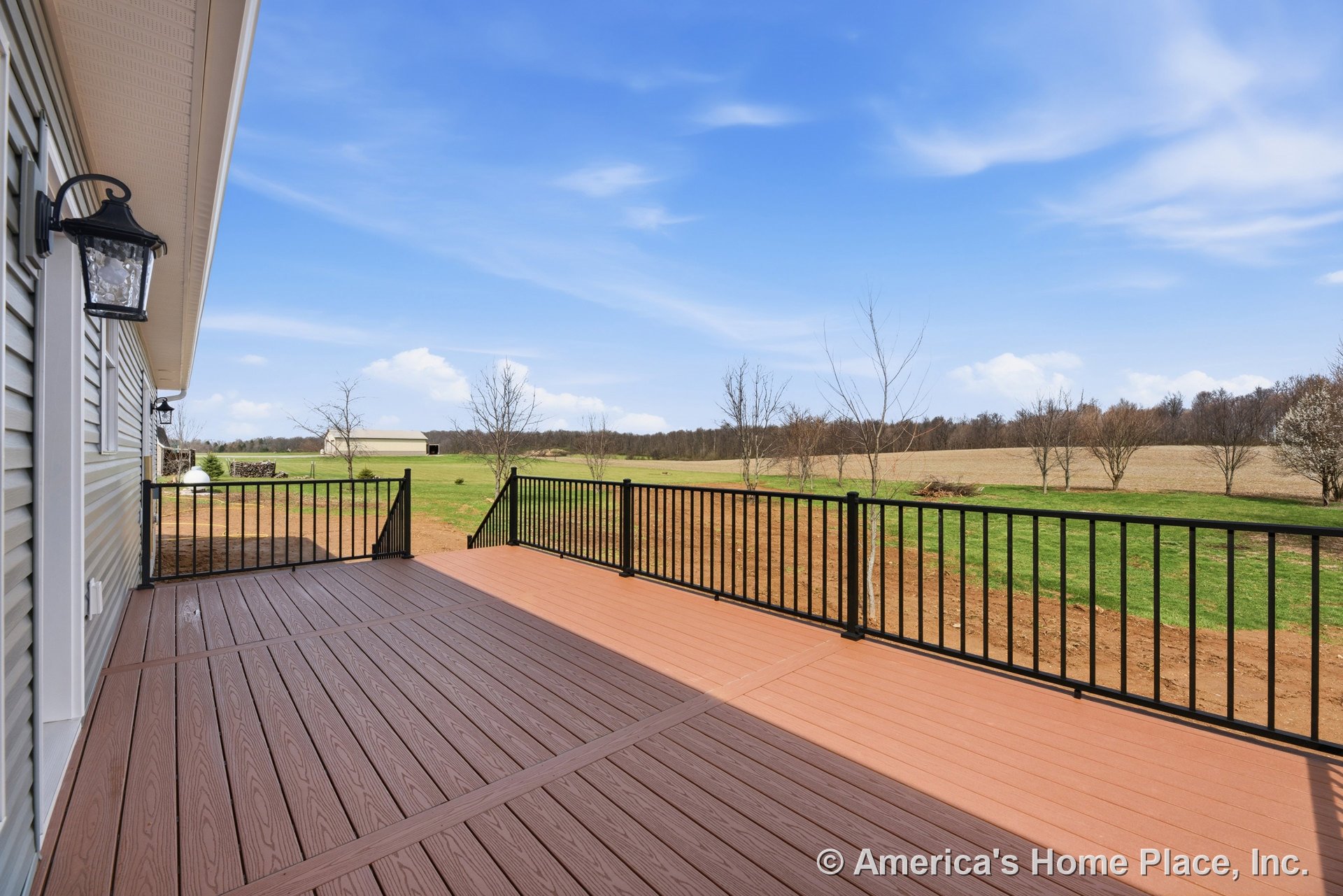 Spacious composite deck with black metal railing, exterior wall siding, outdoor wall lanterns, deck steps leading to yard, overlooking open rural landscape.