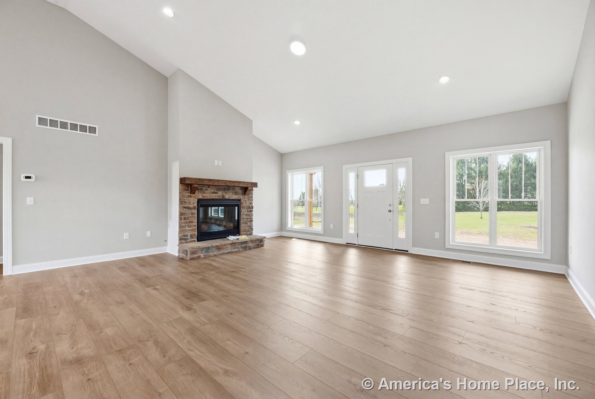 Vaulted ceiling living room with expansive window wall, stone fireplace topped by wood mantel, light wood flooring, white trim, recessed lighting, and open layout.