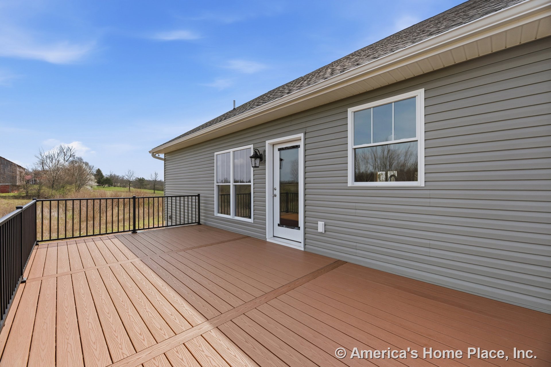 Spacious composite deck with black metal railing, gray vinyl siding, white-trimmed windows, glass exterior door, and outdoor wall-mounted light fixture.