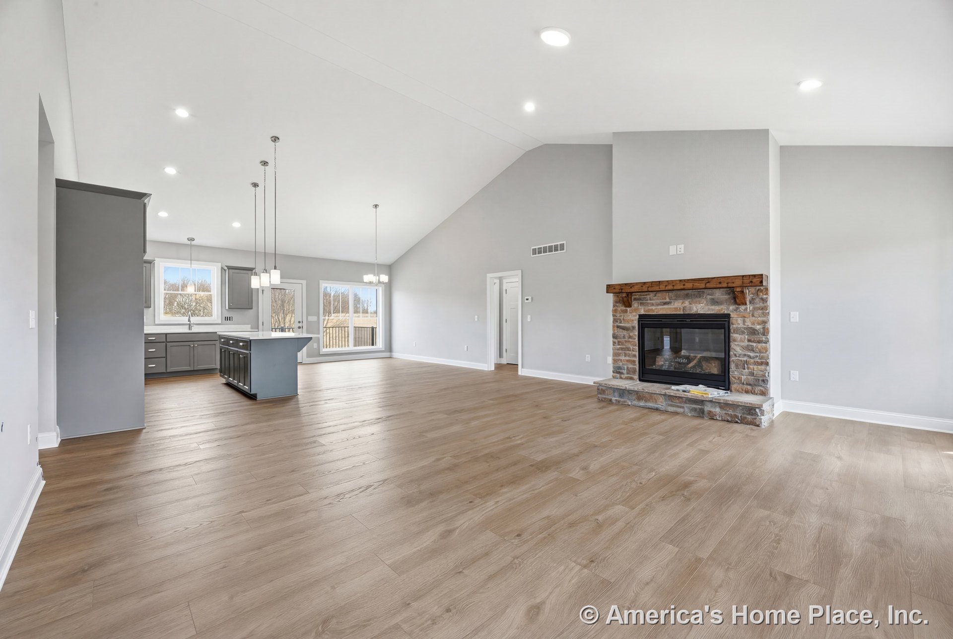 Vaulted ceiling spans open living area with stone fireplace and wood mantel, light wood plank flooring, gray kitchen cabinetry, kitchen island, large window wall, recessed and