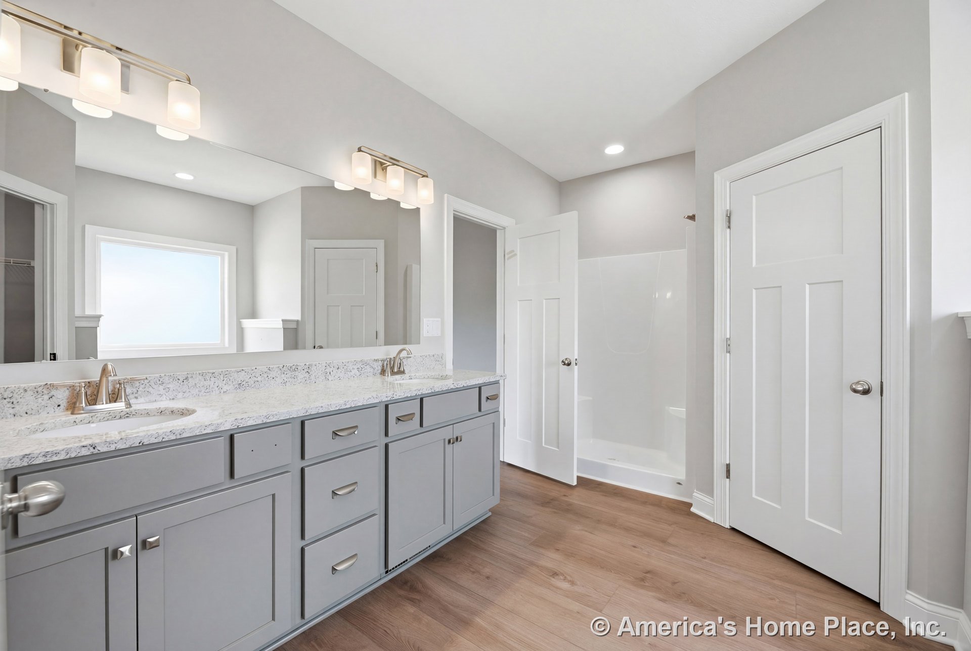 Double sink vanity with gray shaker cabinets and quartz countertop, large wall mirror with built-in lighting fixtures, walk-in shower enclosure, wood flooring, white trim, interior