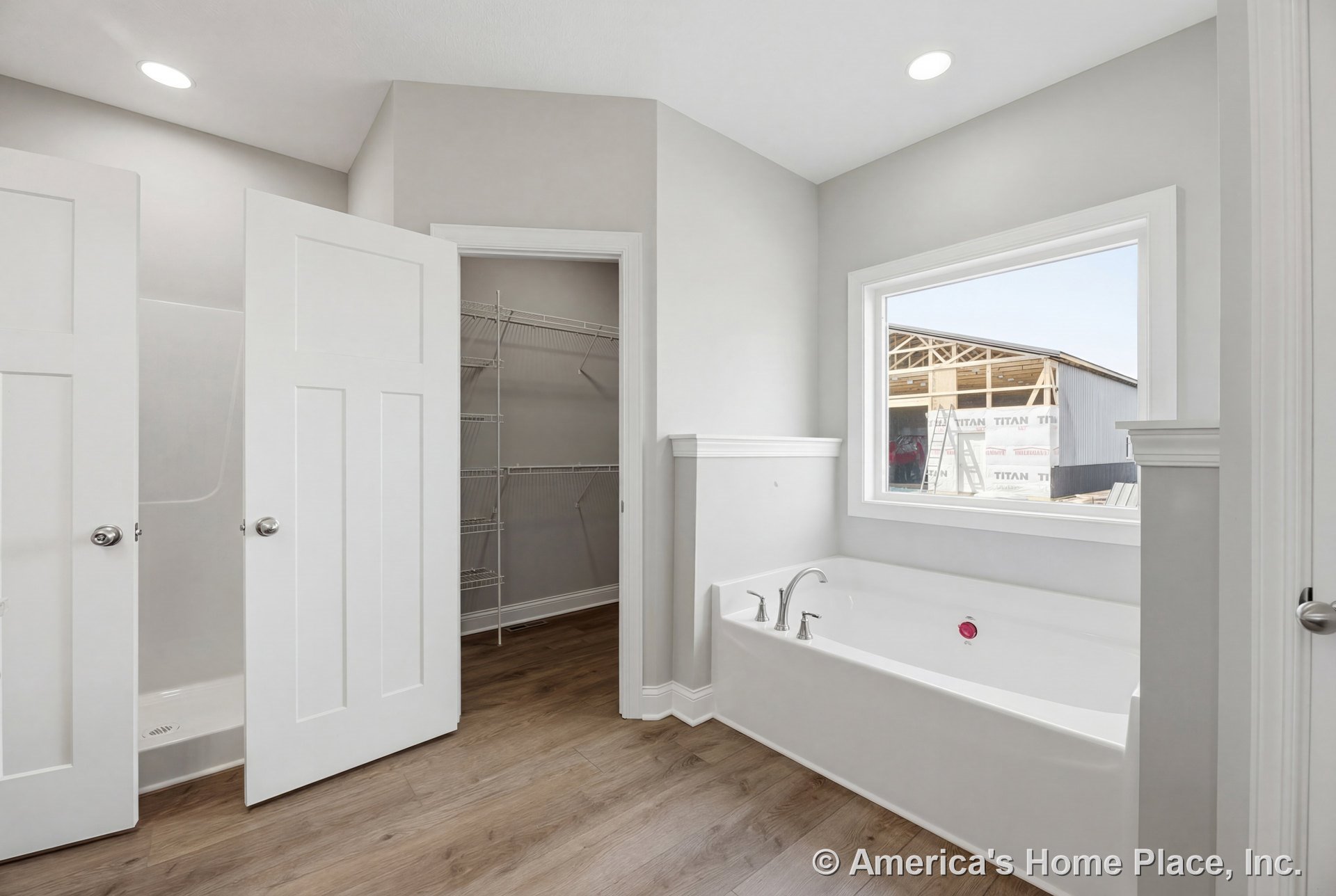 Built-in soaking tub with chrome fixtures beside a large window with white trim, walk-in shower behind a paneled door, closet featuring wire shelving, recessed ceiling lights, and
