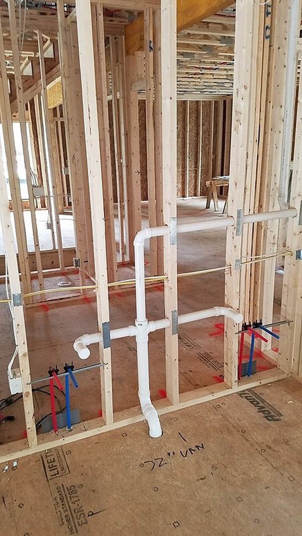 Exposed wood framing, insulation, and plumbing pipes in an unfinished interior with a ladder and construction materials scattered on the floor