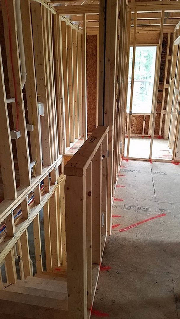 Wood-framed room under construction with exposed beams, concrete floor marked with red lines, window set in brick wall, and visible insulation.