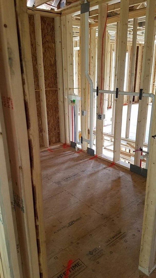 Unfinished room with exposed wood beams, plumbing pipes, building insulation, and red tape marking lines on the subfloor