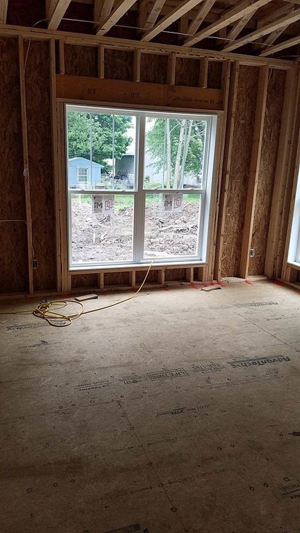 Unfinished room with exposed wood beams, large window overlooking blue house and trees, yellow wire or hose lying on subfloor