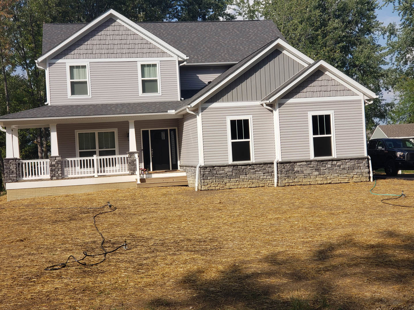 Two-story home with light siding, black-framed windows, black front door with white trim, covered porch, grassy yard, mature trees in background, black truck parked in driveway