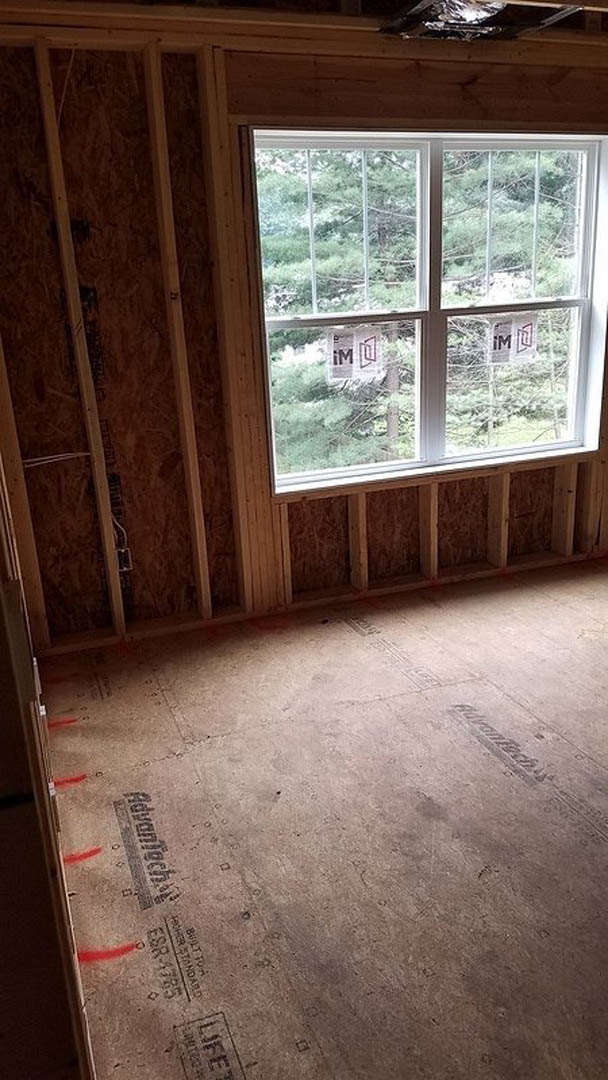 Sunlit room featuring wide plank wood flooring, large window with view of leafy trees, white walls, and minimalist trim.
