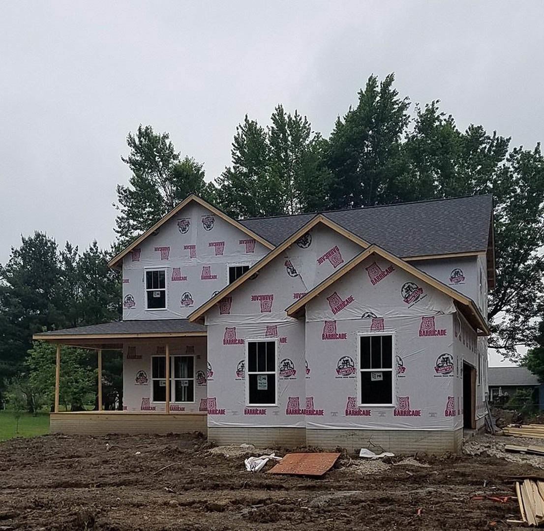 Framed house under construction with plastic covering, brown wood siding, pile of lumber on ground, white sign taped to window, trees in background