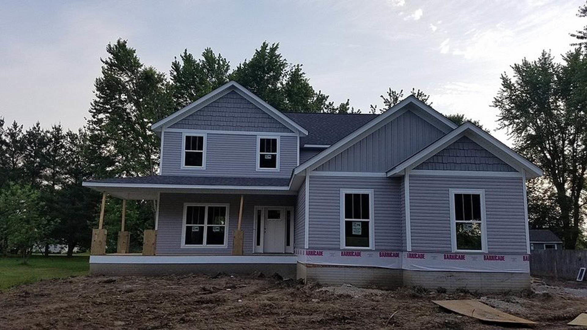 Partially built house with exposed framing, white window and door, red tape around openings, construction sign posted on window, surrounded by trees in background