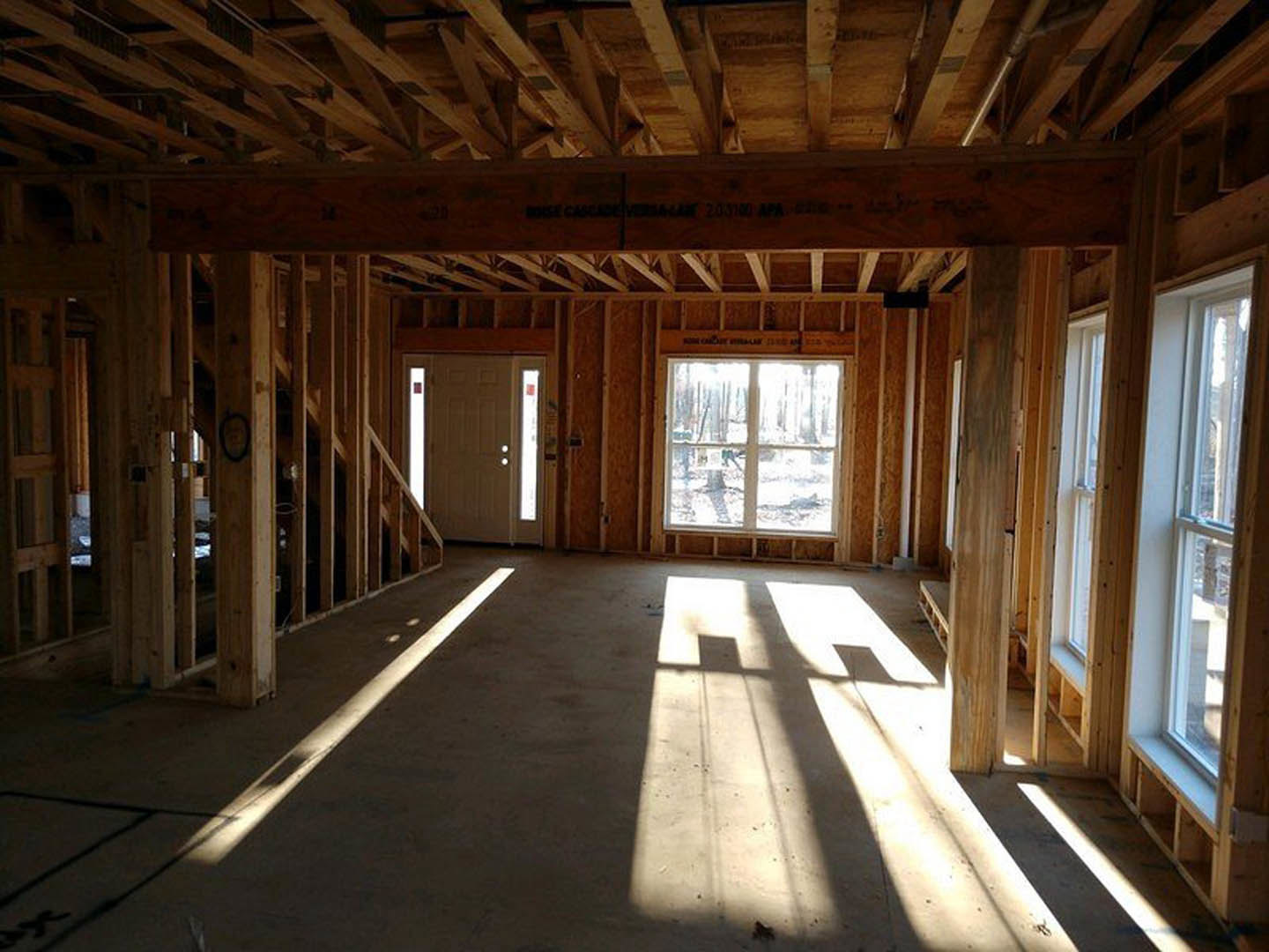 Sunlit room with hardwood floors, white door, large window overlooking forest, wood plank ceiling, and natural light streaming across the floor.