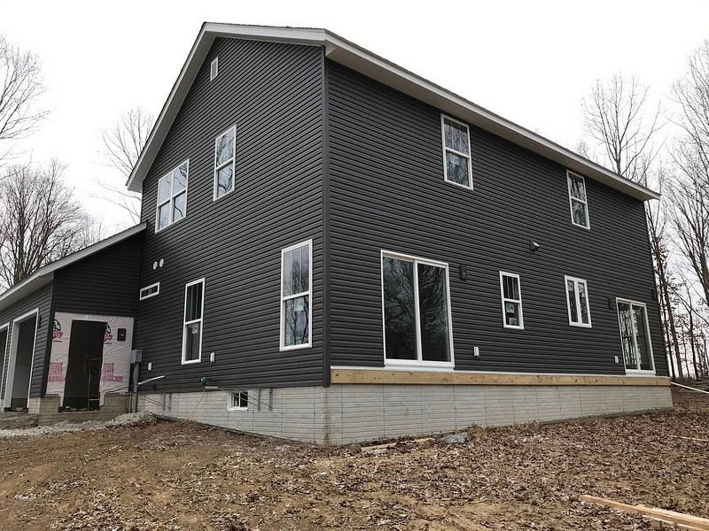 Grey siding house under construction with white framed window, black door, dirt path and scattered leaves in front, trees and sky in background
