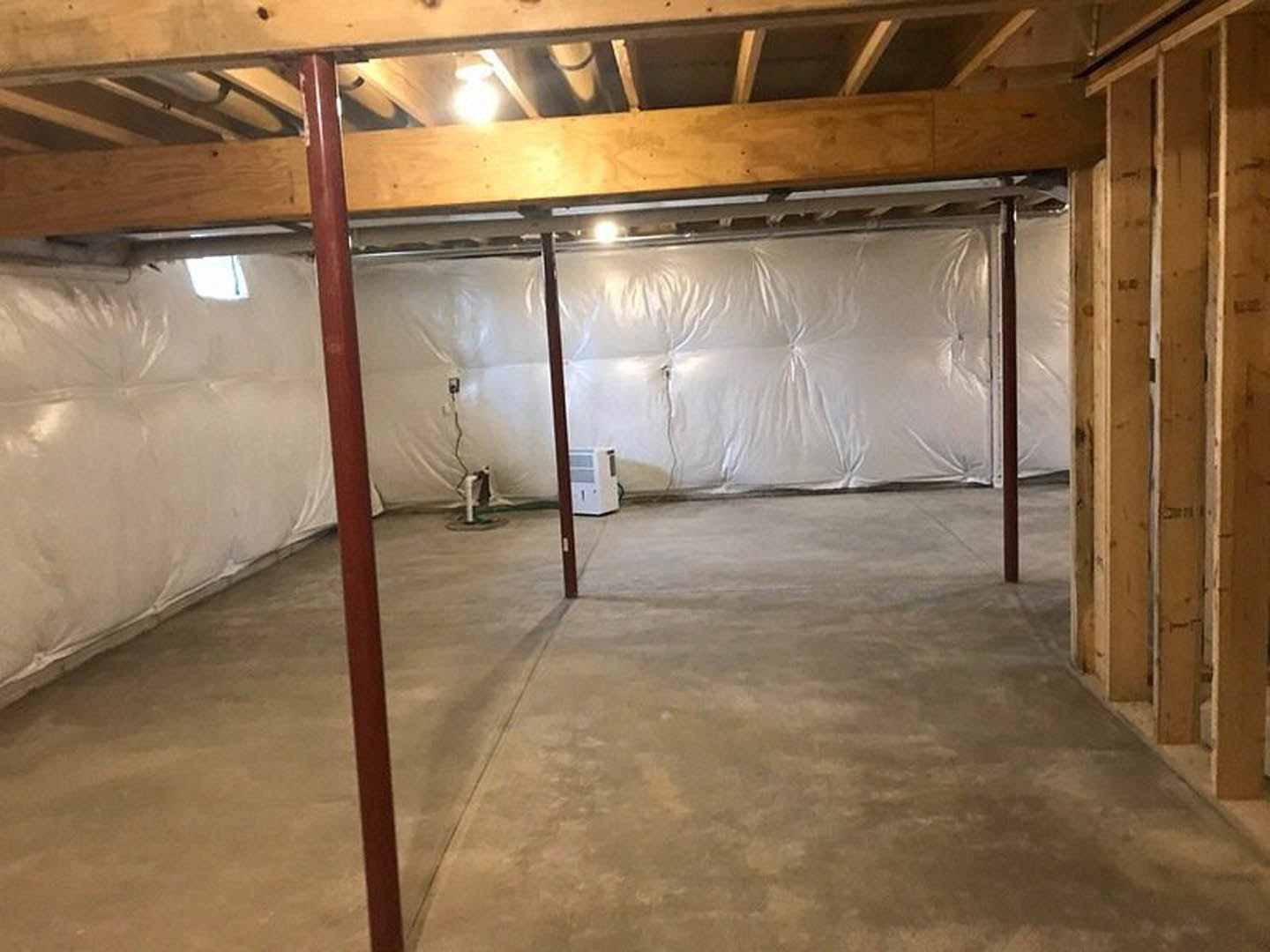Unfinished basement room with exposed wooden beams, white sheet draped over an object, white door with window, red pole with white cover, visible insulation and plaster, light bulb
