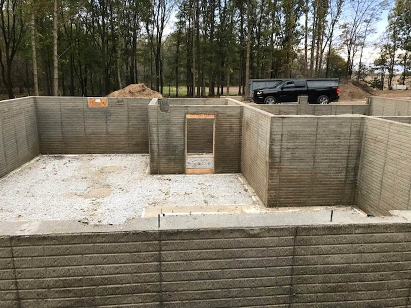 Partially built home with exposed concrete walls, cement foundation, and construction truck parked behind; trees and sky visible in background