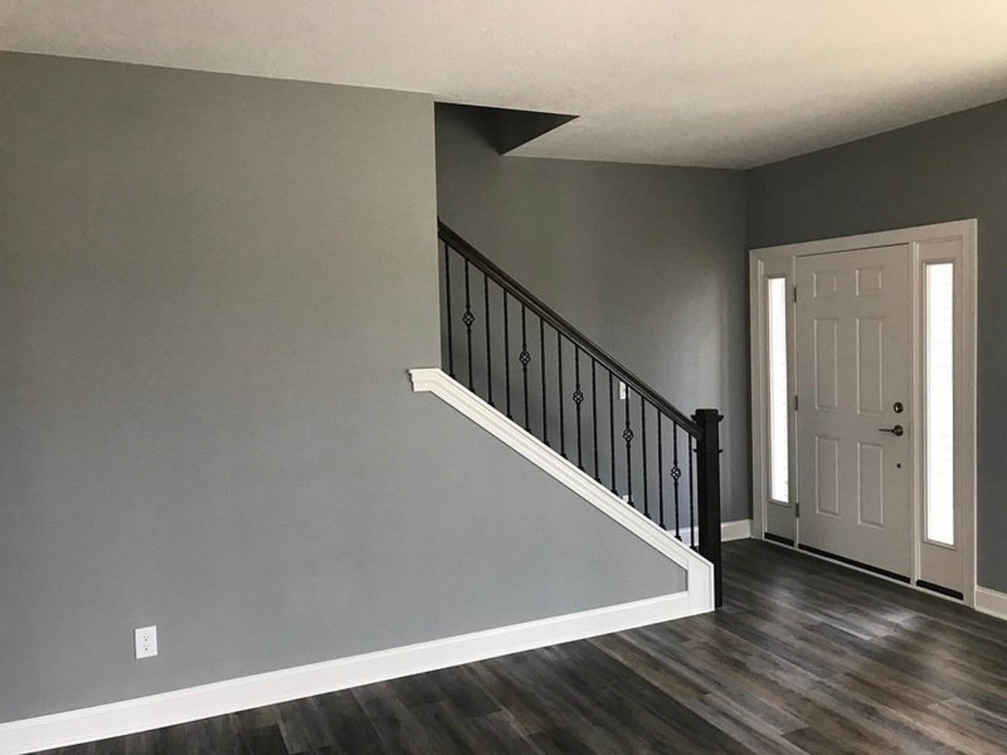 Wood staircase with black metal railing, white walls and baseboards, laminate flooring, white door with silver doorknob, electrical outlet, and ceiling corner visible.
