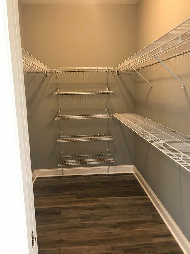 Empty closet with built-in white shelves, wood flooring, white trim, and a metal hanging bar on the wall